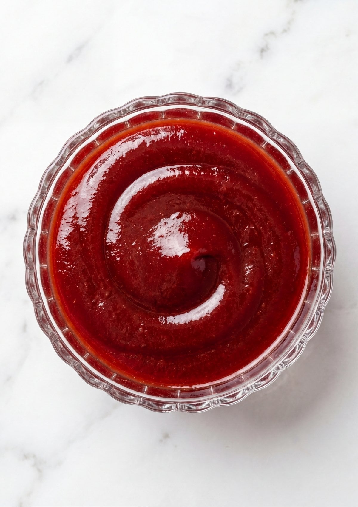A small glass bowl filled with a smooth red ketchup glaze swirled in the center sitting on a white marble background.