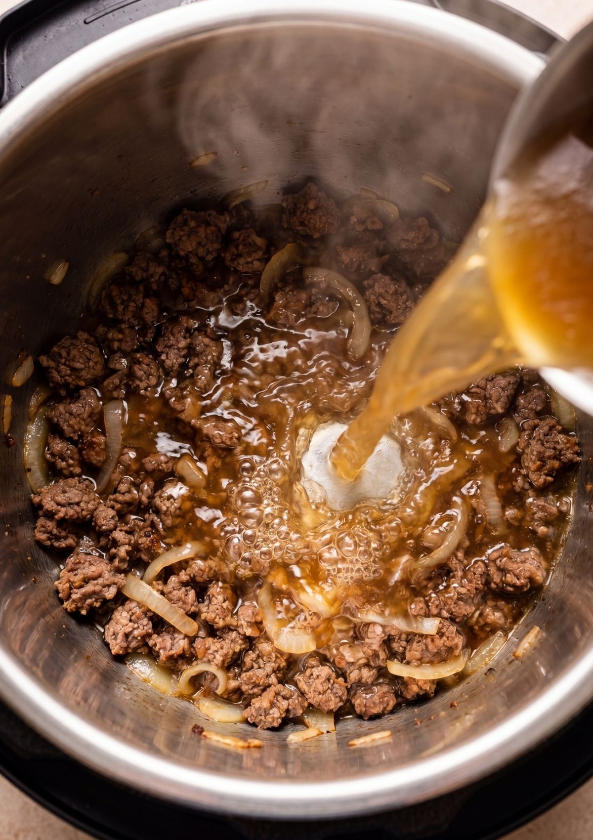 A hand pouring rich beef broth into an Instant Pot containing browned ground beef and sautéed onions to deglaze the bottom.