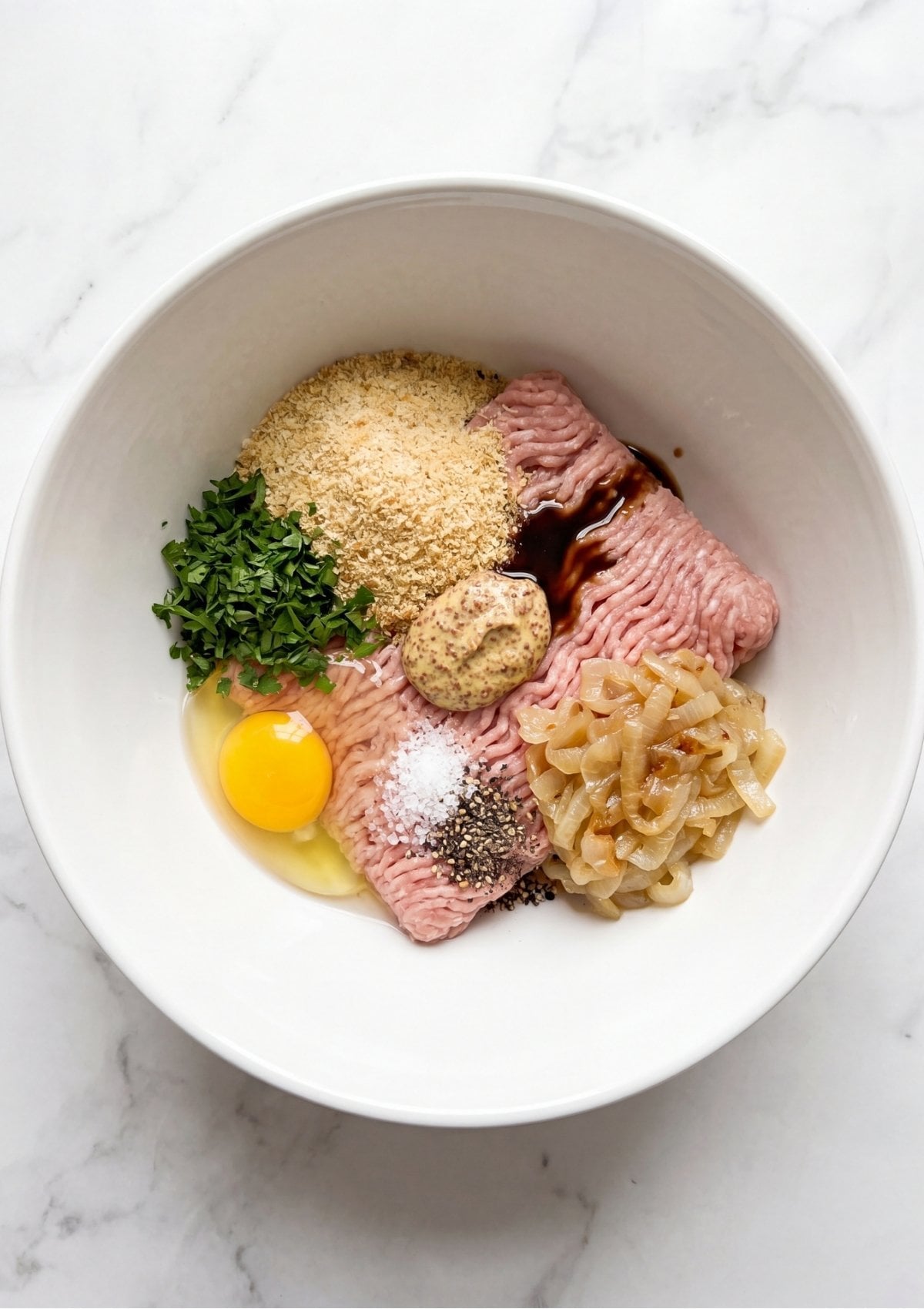 A white mixing bowl filled with raw ground turkey, breadcrumbs, chopped parsley, a raw egg, whole grain mustard, salt, pepper, and sautéed onions before being mixed
