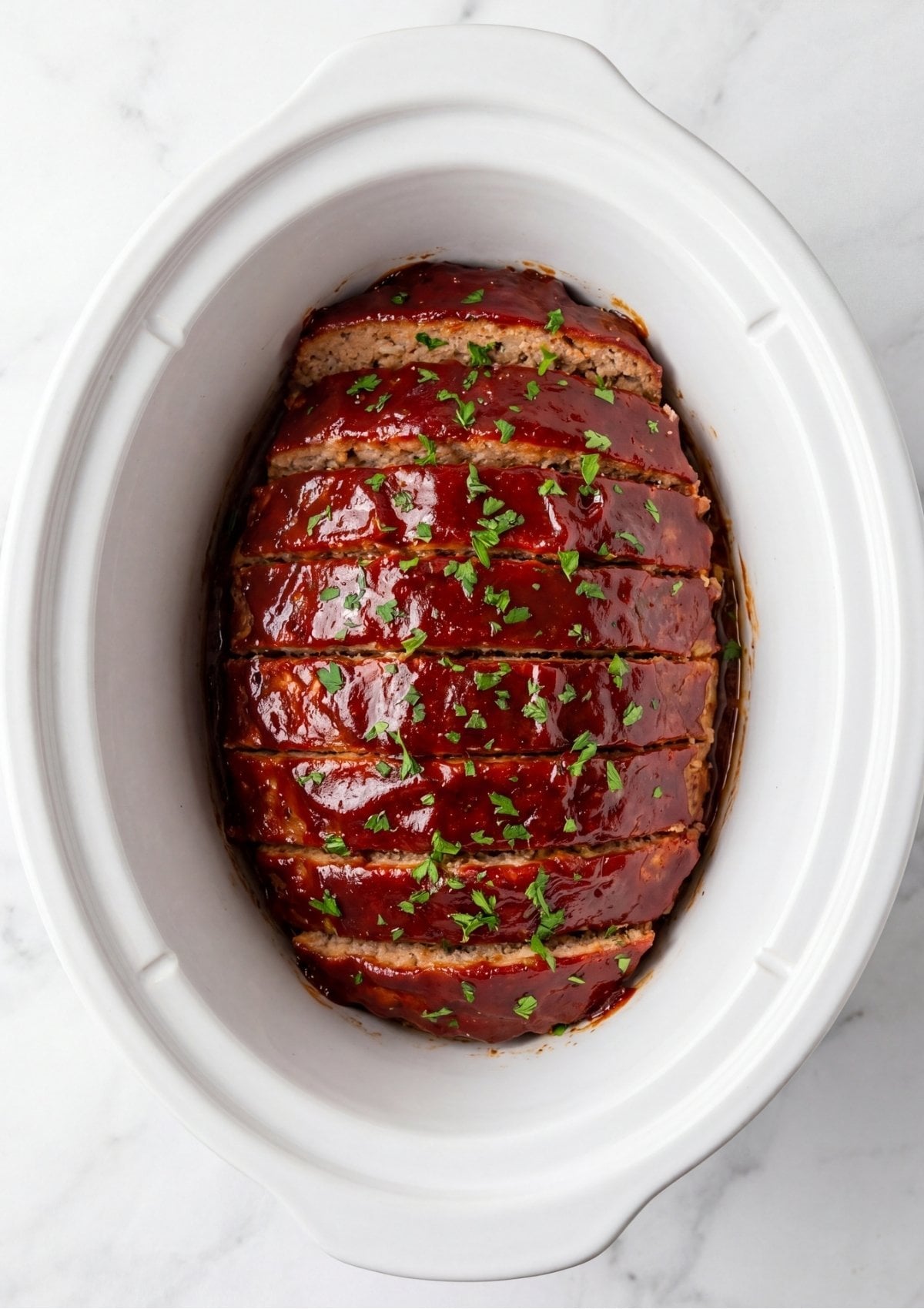 Sliced turkey meatloaf topped with a red ketchup glaze and chopped fresh parsley inside a white ceramic crockpot bowl