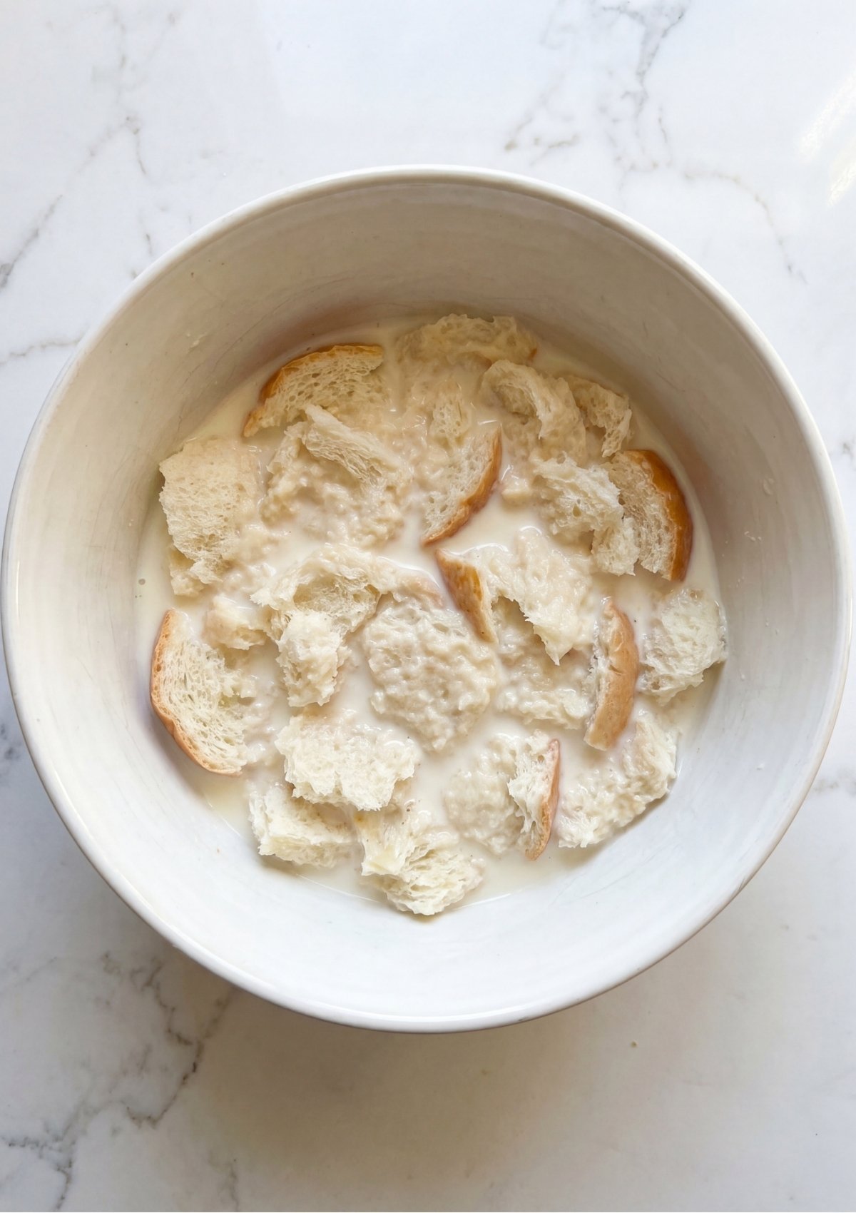 White bread pieces soaking in a small pool of milk inside a large white ceramic mixing bowl