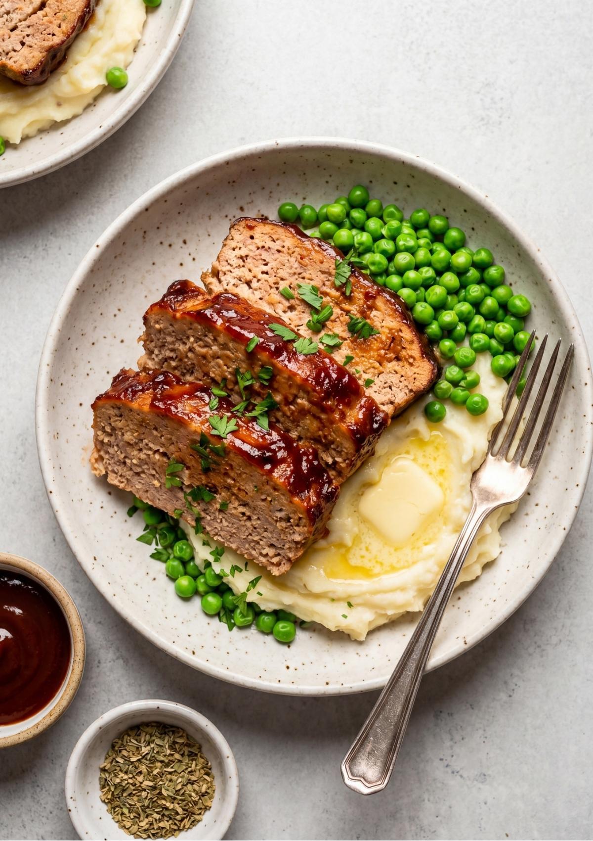 Three thick slices of glazed turkey meatloaf arranged on top of creamy mashed potatoes with a side of green peas and a fork.