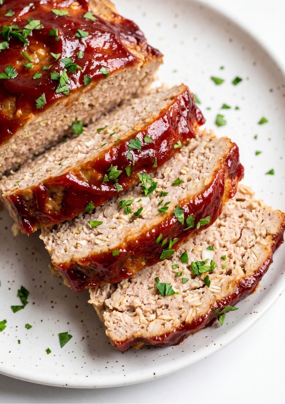 Close-up of several thick slices of turkey meatloaf showing a visible texture of oats and onions, topped with a red glaze and fresh parsley on a speckled white plate.