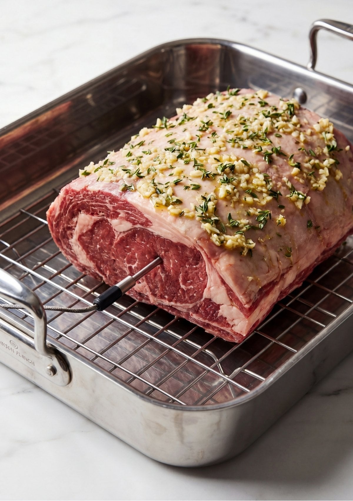 Raw beef roast rubbed with minced garlic and fresh thyme sitting on a metal wire rack inside a roasting pan with a probe thermometer inserted