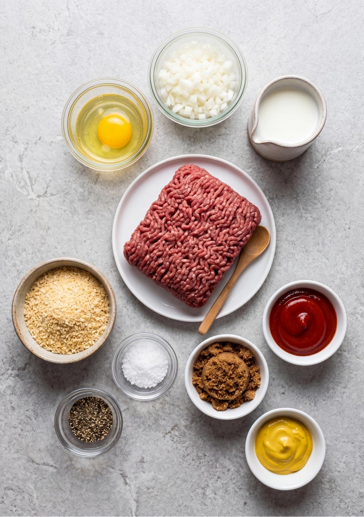 A flat-lay view of ingredients including ground beef, breadcrumbs, an egg, chopped onions, milk, ketchup, mustard, brown sugar, salt, and pepper.