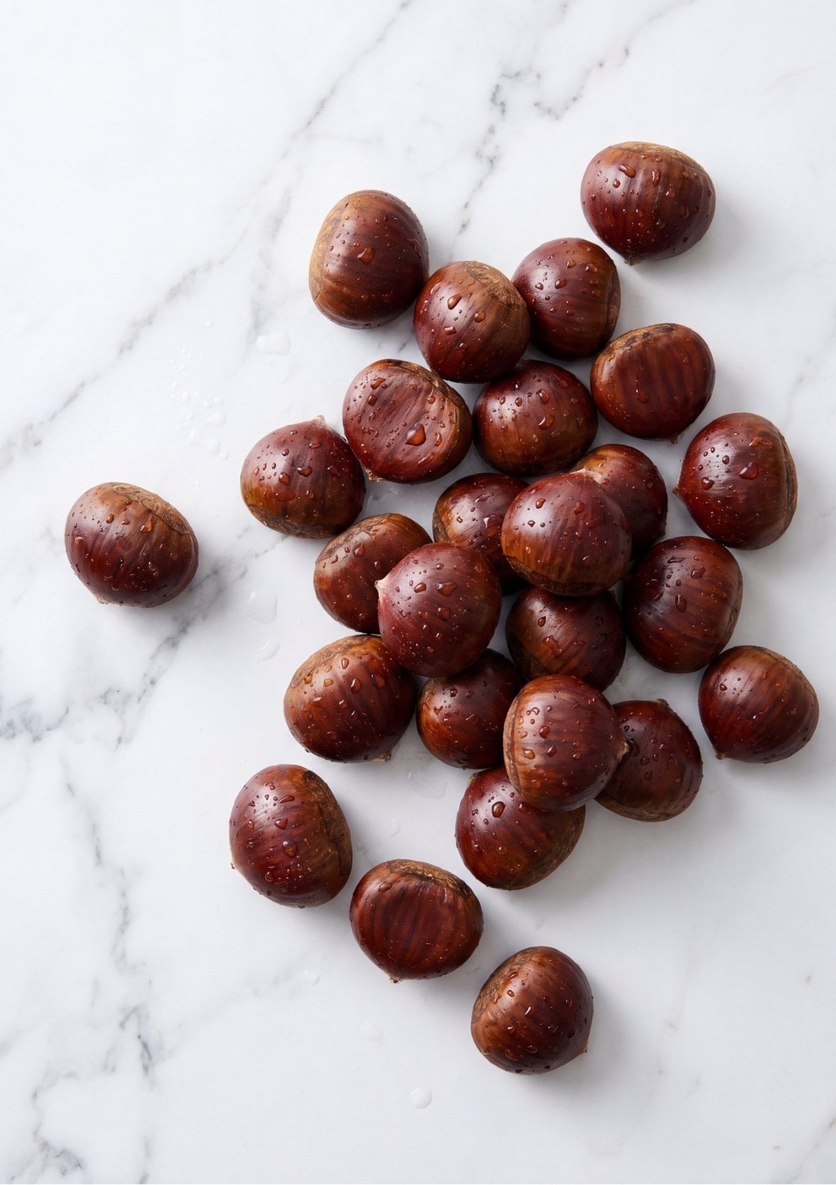 A cluster of fresh, wet whole chestnuts with shiny brown shells resting on a white marble countertop.