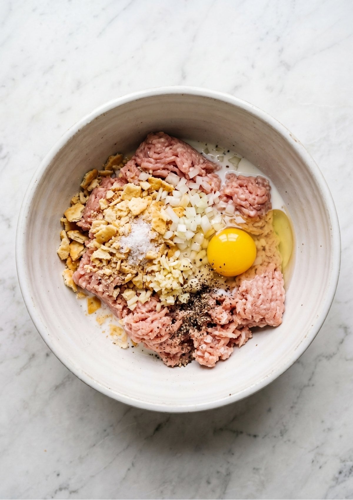A white ceramic bowl containing the ground turkey combined with the egg, onions, garlic, and cracker crumbs before being mixed.