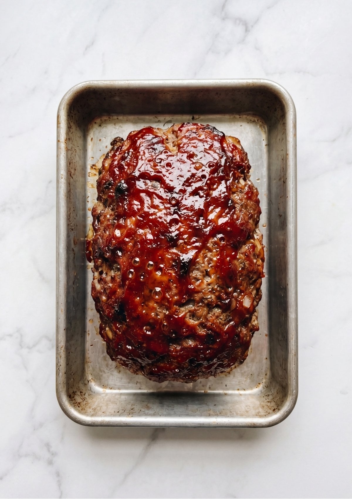 A whole, cooked meatloaf covered in a bubbling, caramelized glaze sitting in a rectangular metal baking pan.