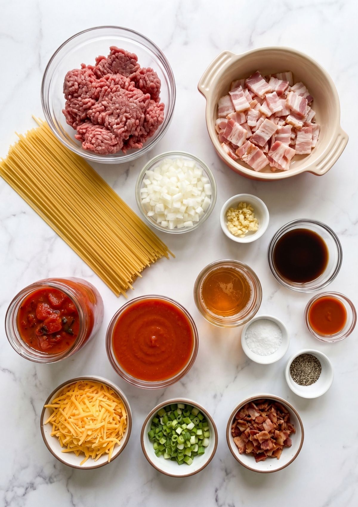 A flat lay of spaghetti noodles, ground beef, raw bacon, chopped onions, garlic, and sauces in glass bowls on white marble.