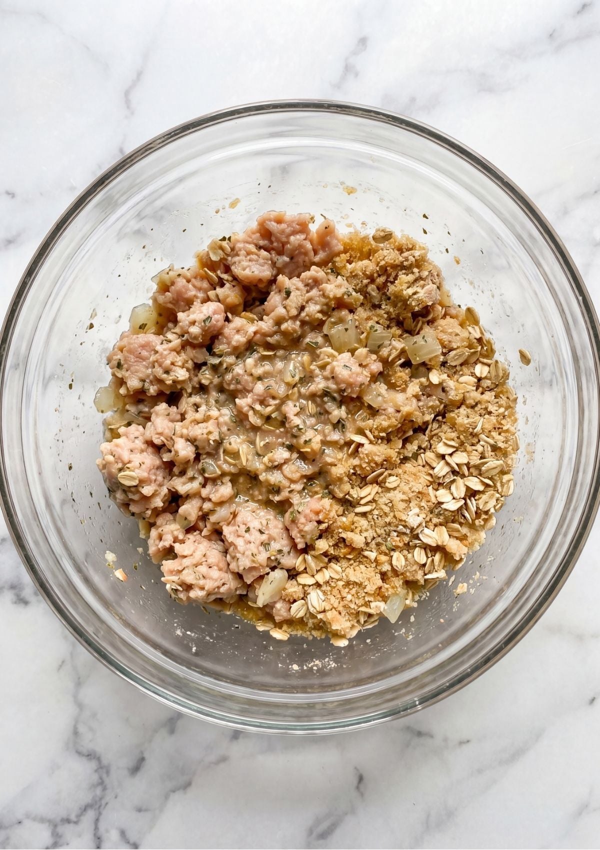 Close-up of ground chicken being combined with oats and breadcrumbs in a glass bowl on a white marble countertop.
