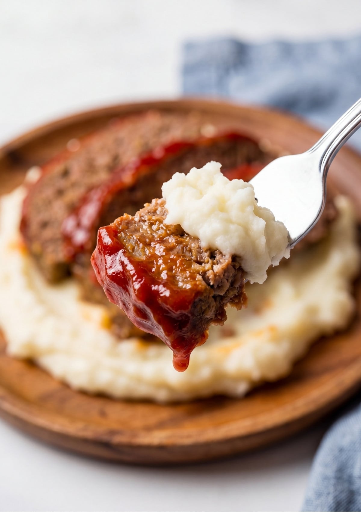 A silver fork holding a piece of glazed meatloaf topped with a dollop of creamy mashed potatoes over a wooden plate