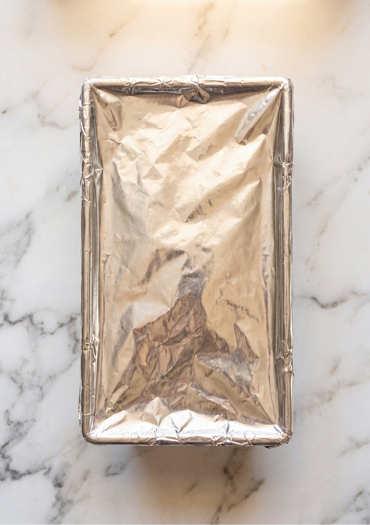 A top down view of a rectangular loaf pan covered tightly with silver aluminum foil on a white marble surface