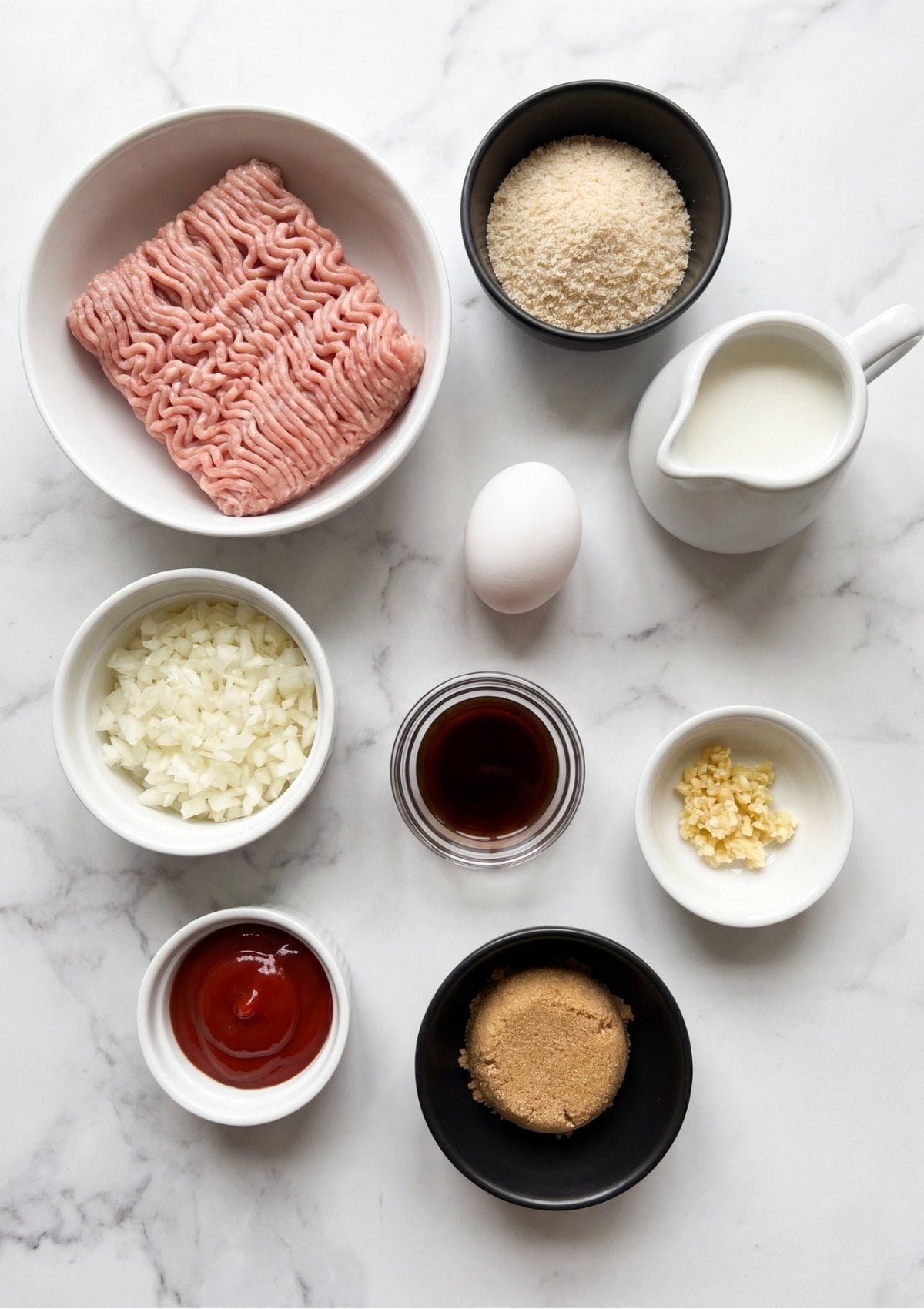 A flat lay view of recipe ingredients including ground turkey, breadcrumbs, diced onions, milk, an egg, garlic, ketchup, and brown sugar in small bowls.
