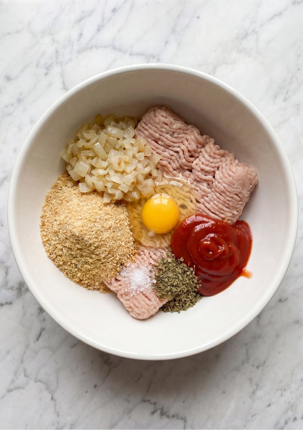 Top-down view of unmixed meatloaf ingredients including raw ground turkey, a whole egg yolk, breadcrumbs, ketchup, sautéed onions, dried marjoram, and salt neatly arranged inside a white ceramic bowl