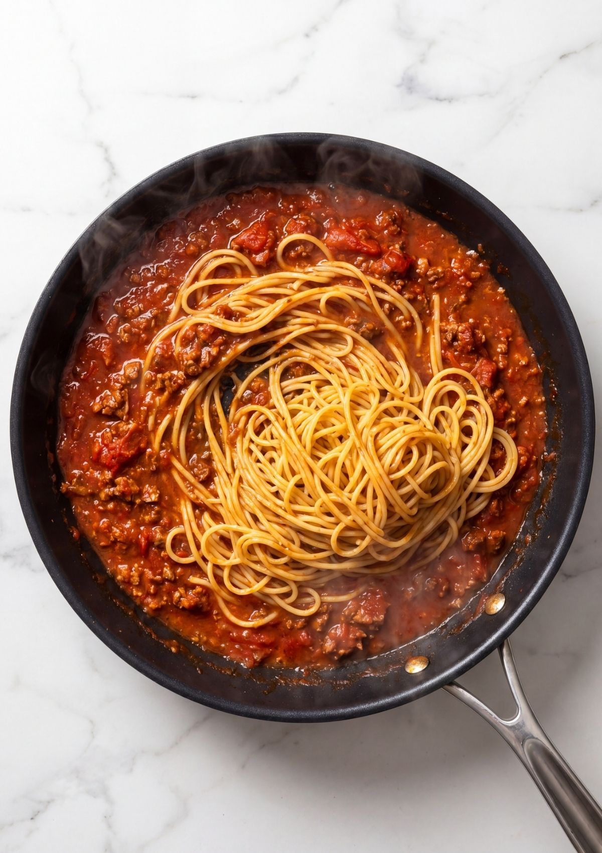 Long spaghetti noodles being added to a simmering meat and tomato sauce in a black pan on a white marble background.