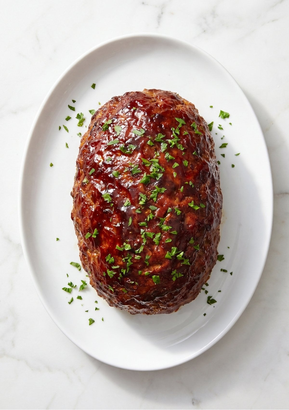 A whole cooked meatloaf topped with a shiny red glaze and sprinkled with fresh parsley on a white oval platter