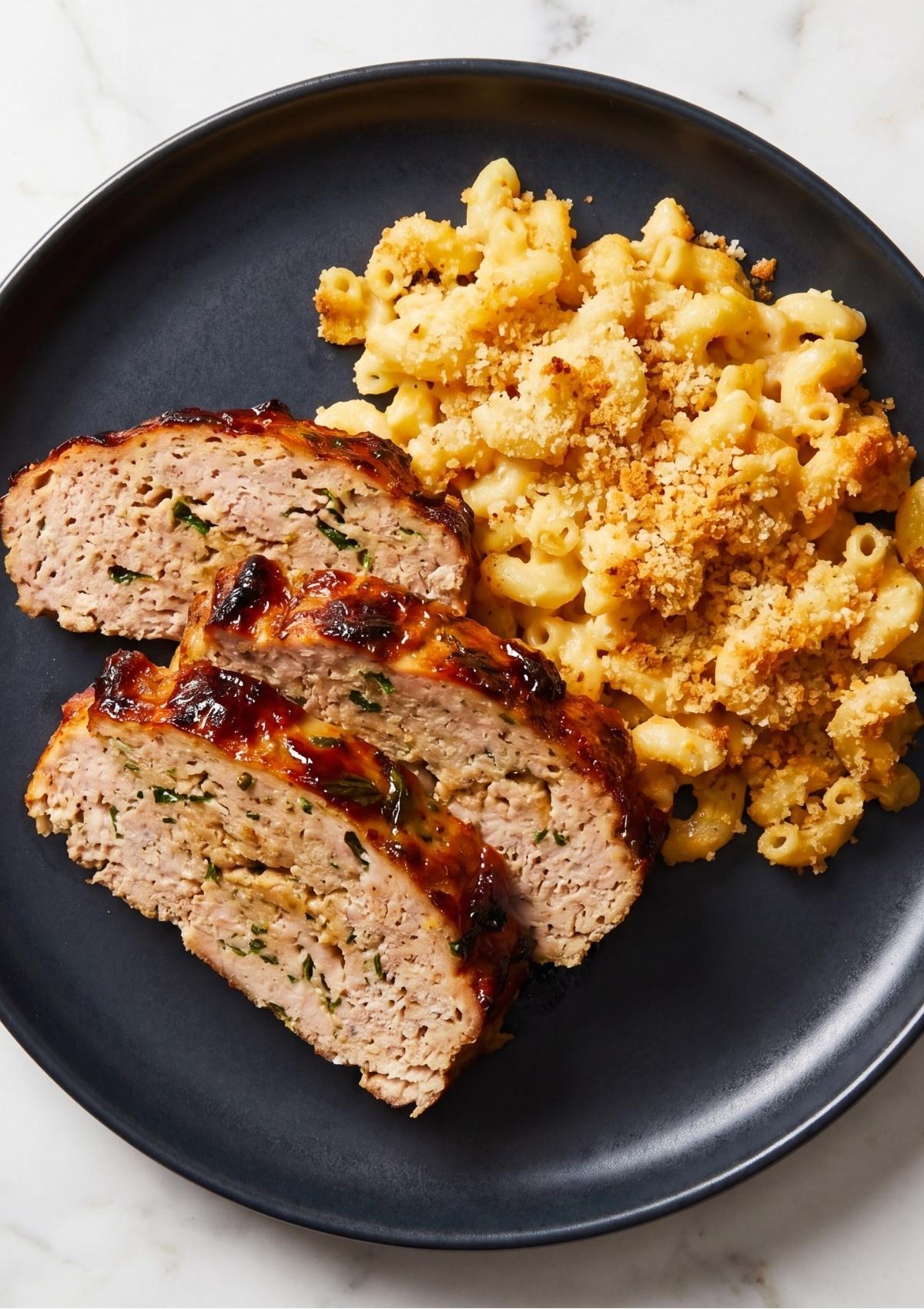 Three thick slices of glazed turkey meatloaf served alongside a generous portion of creamy macaroni and cheese with a breadcrumb crust on a dark blue plate