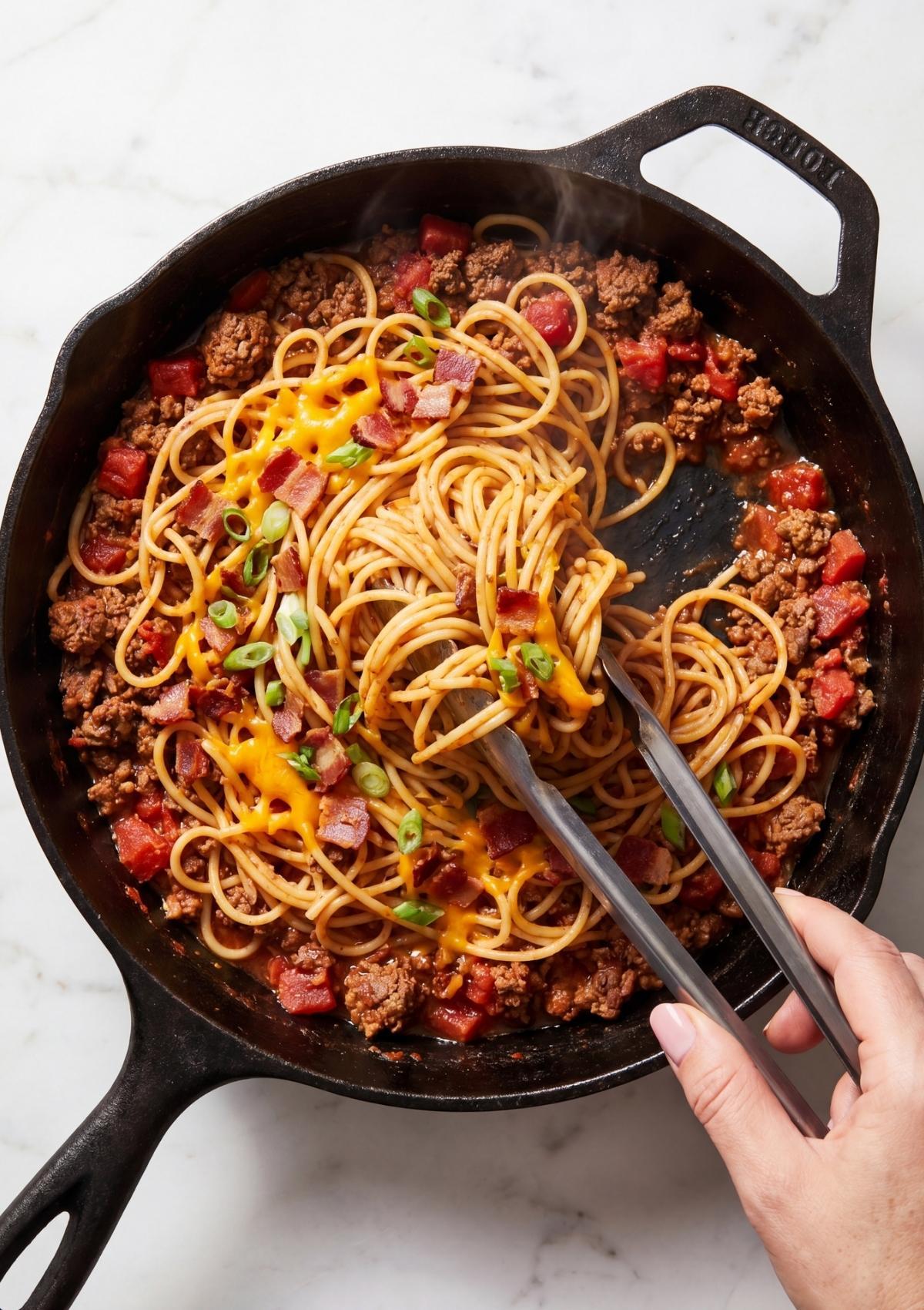 A woman with natural nails using metal tongs to toss spaghetti with bacon, cheese, and green onions in a cast iron skillet.