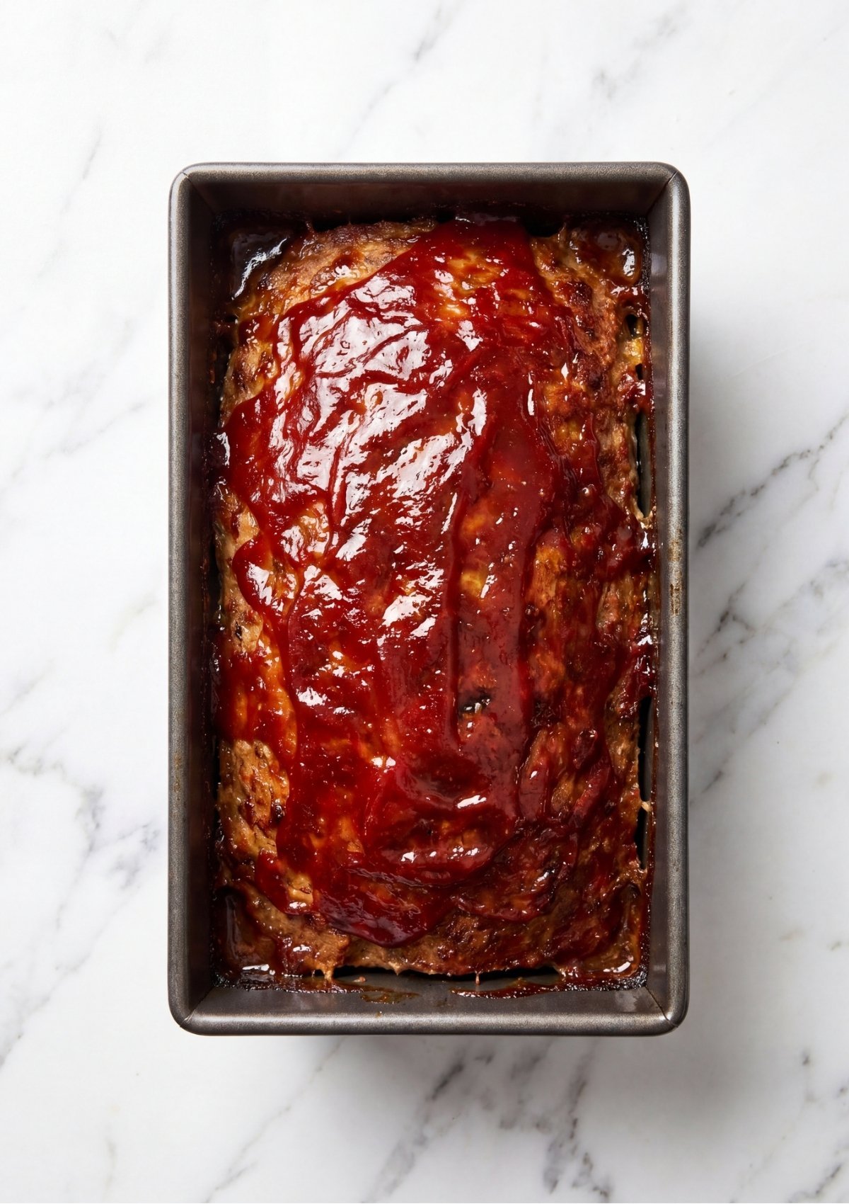 A glossy red ketchup glaze spread evenly across the top of a meatloaf inside a metal loaf pan before baking.