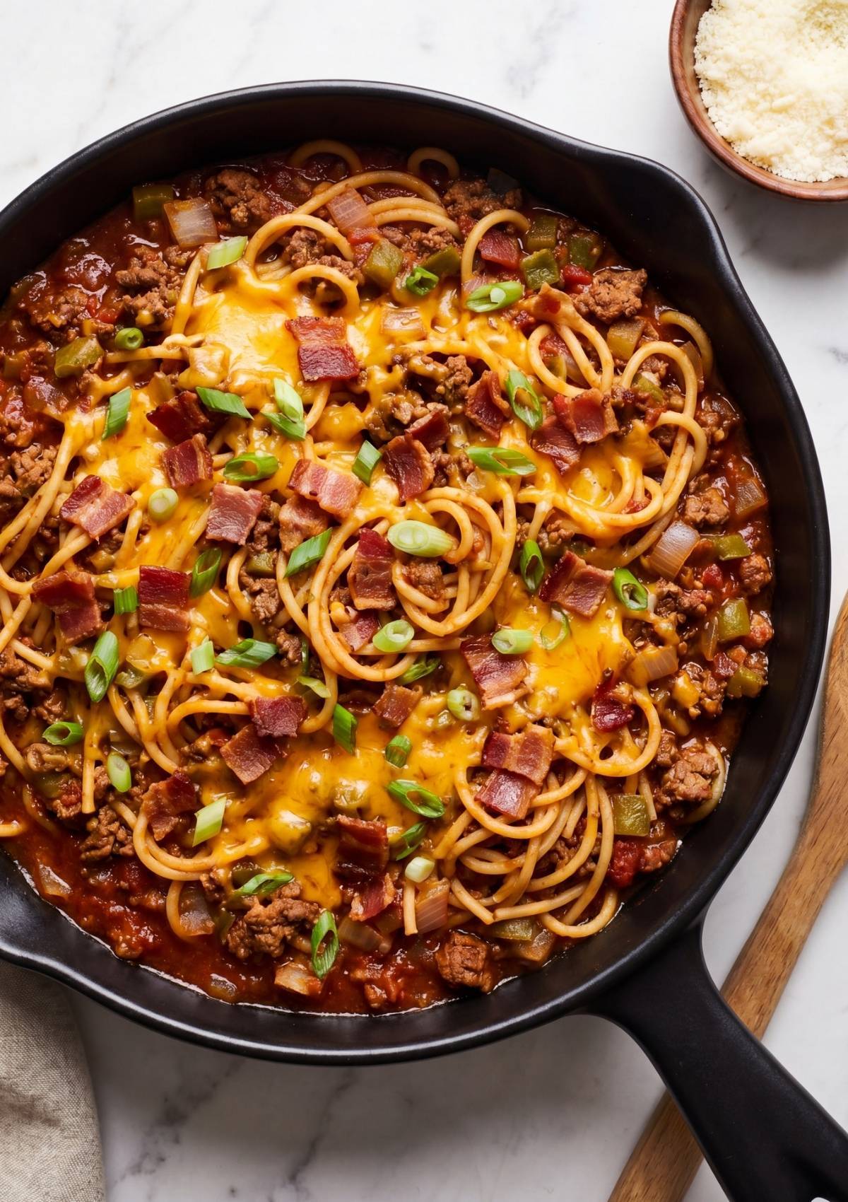 A top-down shot of high-protein cowboy spaghetti in a cast iron pan, garnished with plenty of melted cheddar, bacon bits, and scallions.