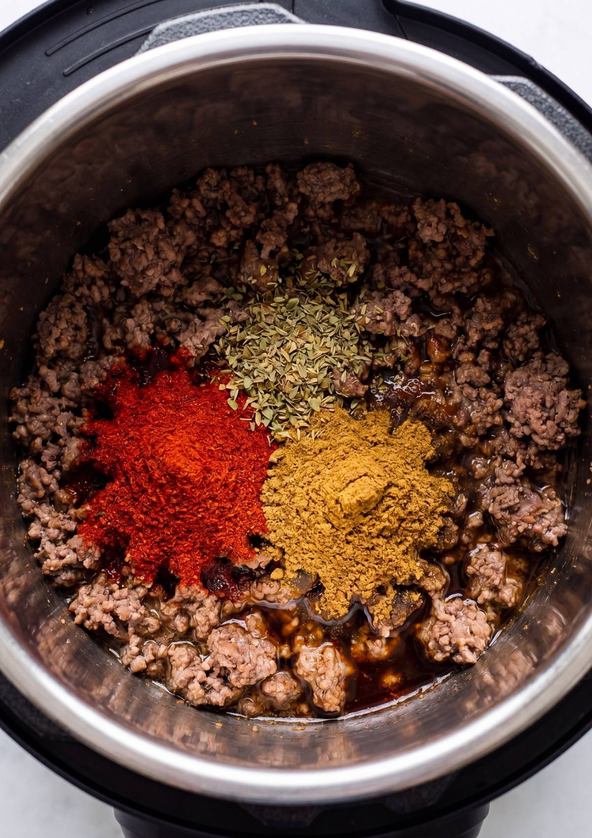 Chili powder, cumin, and dried oregano being added to the browned beef and aromatics inside the Instant Pot.