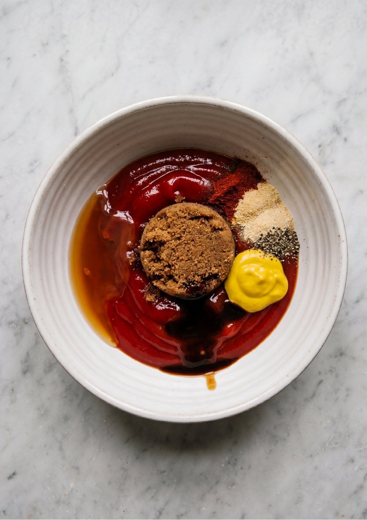 A white bowl containing ketchup, brown sugar, mustard, spices, and liquid ready to be mixed into a Meatloaf Glaze Recipe.