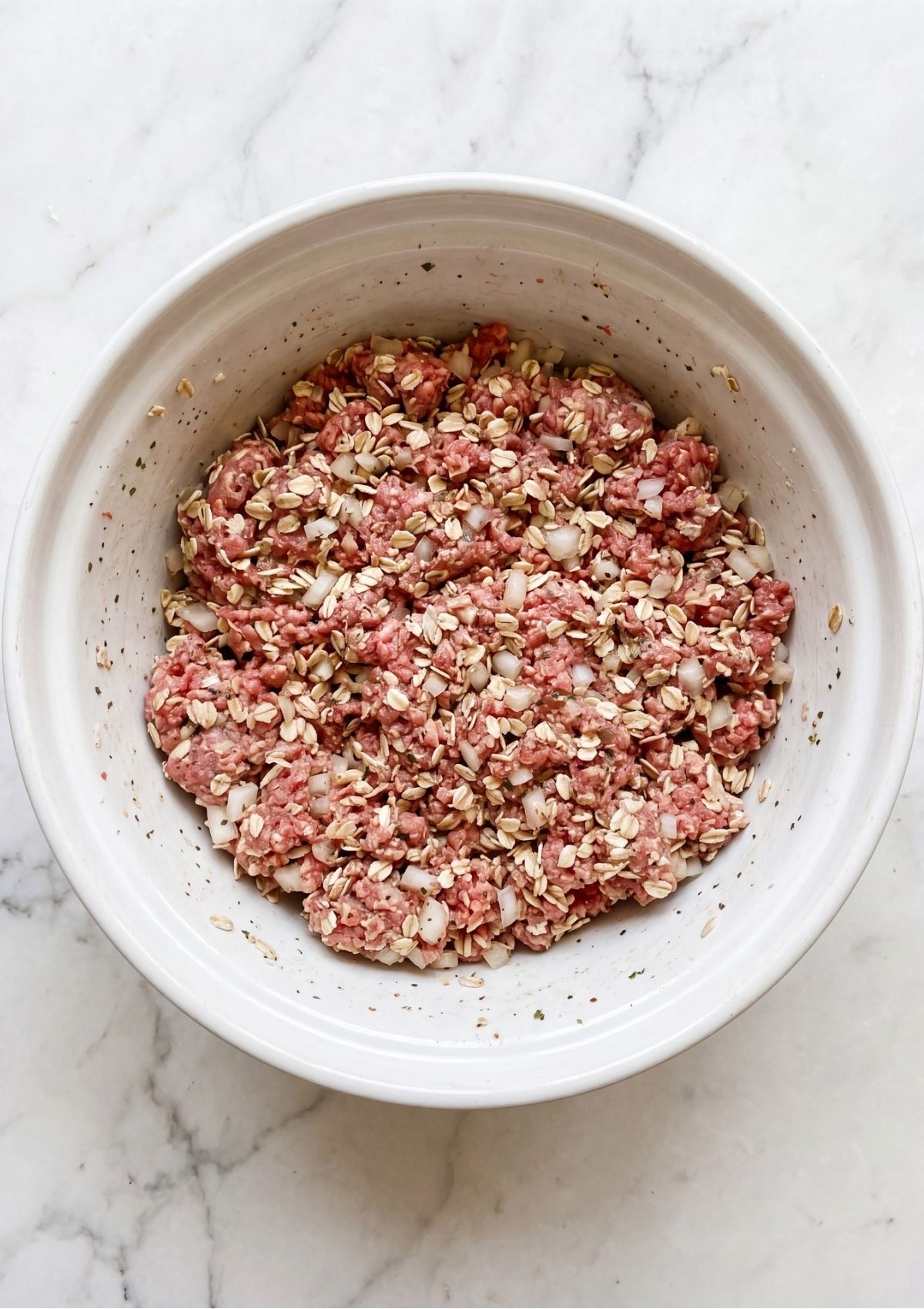 Raw ground beef combined with rolled oats and onions in a white mixing bowl.