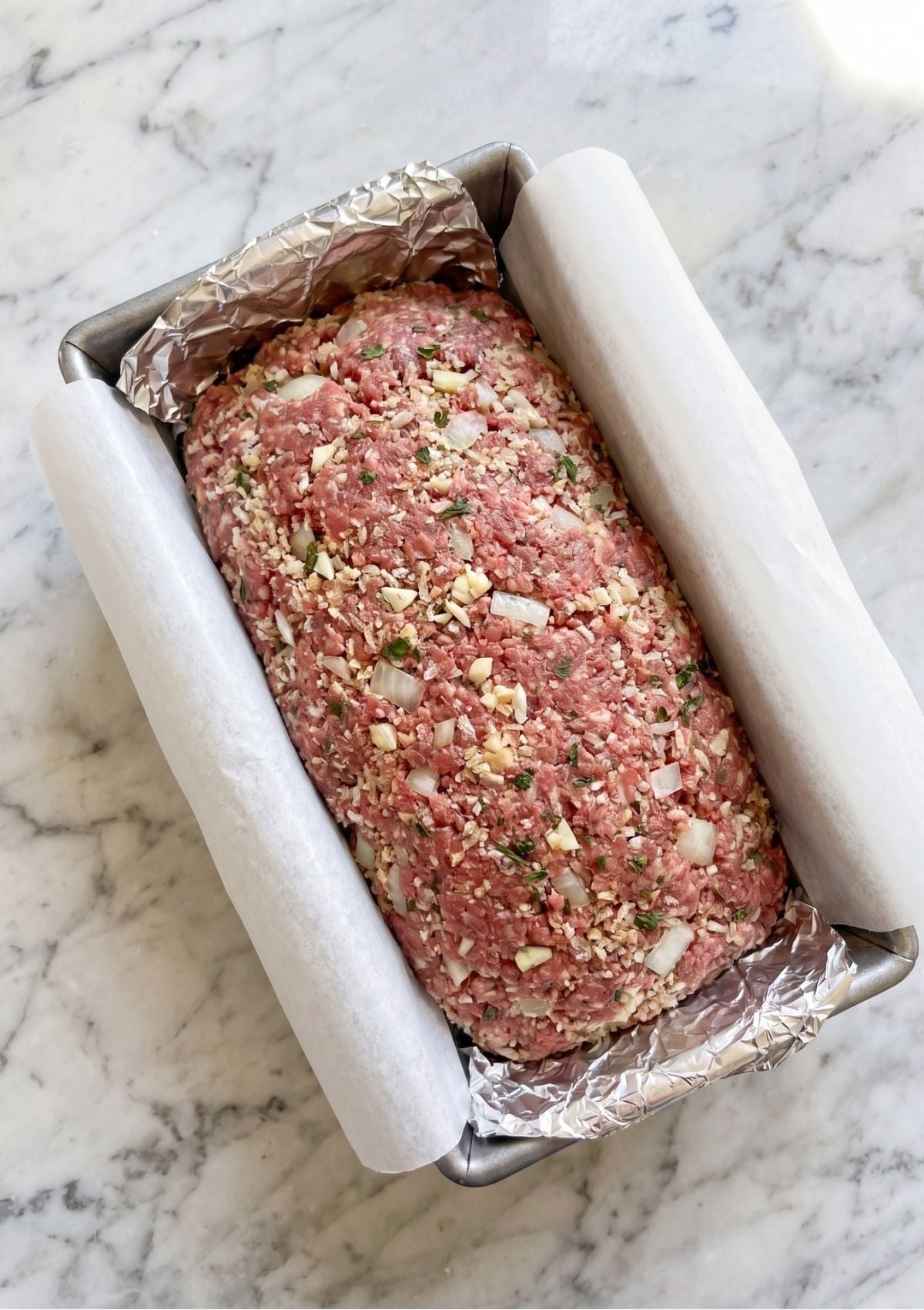A raw meatloaf mixture shaped into a loaf pan lined with parchment paper and foil, ready to be baked on a marble countertop.