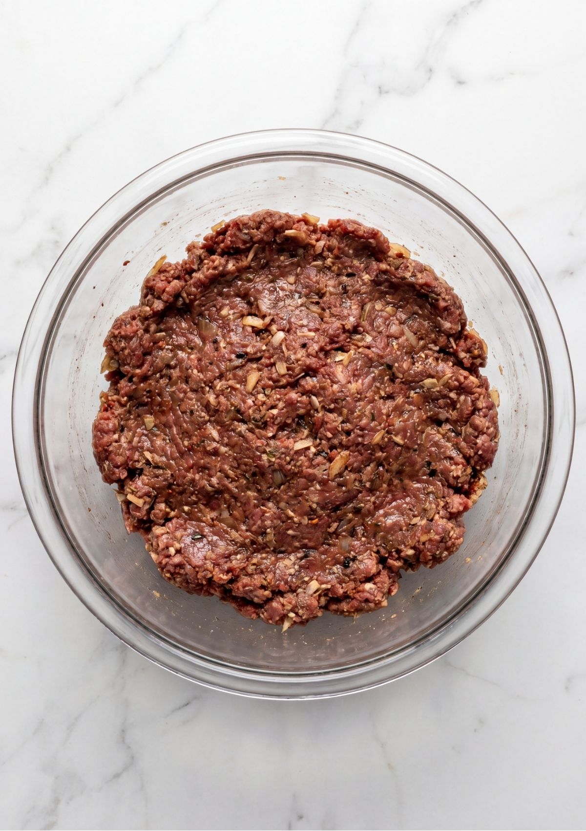 A large clear glass bowl filled with the raw meatloaf mixture, showing ground beef thoroughly combined with onions, seasonings, and binders ready for shaping.