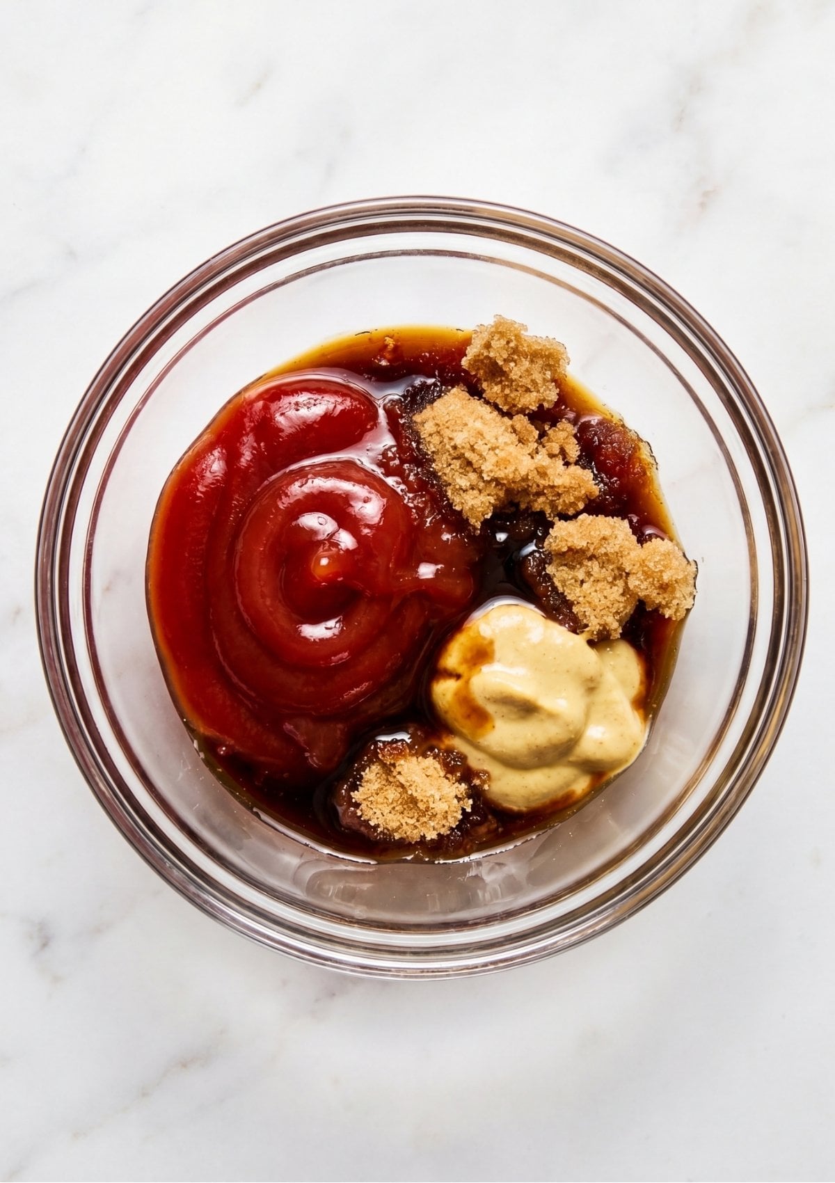 A clear glass bowl containing ketchup, brown sugar, Dijon mustard, and Worcestershire sauce before being mixed.