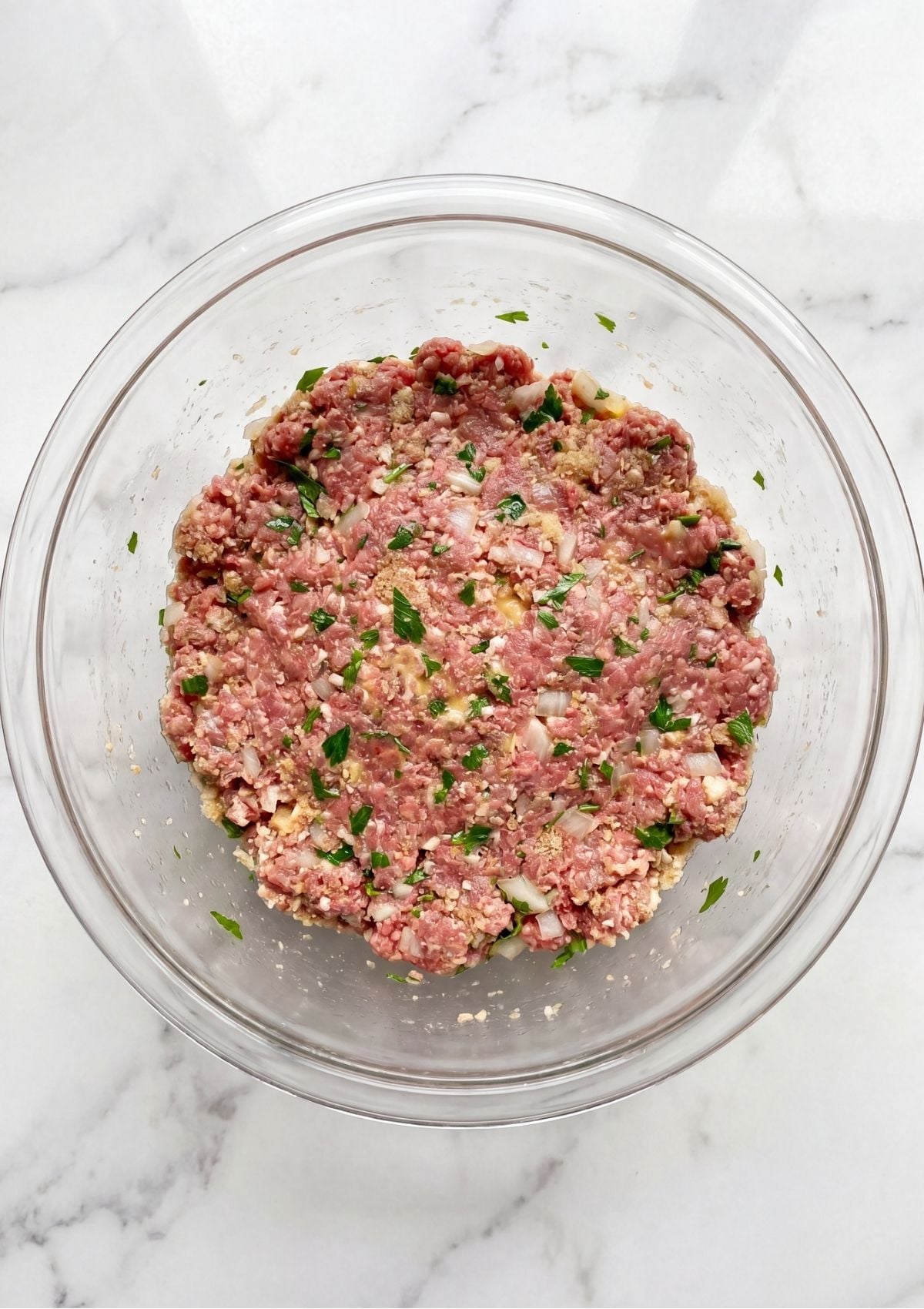 Raw ground beef mixture with chopped onions and parsley inside a clear glass mixing bowl