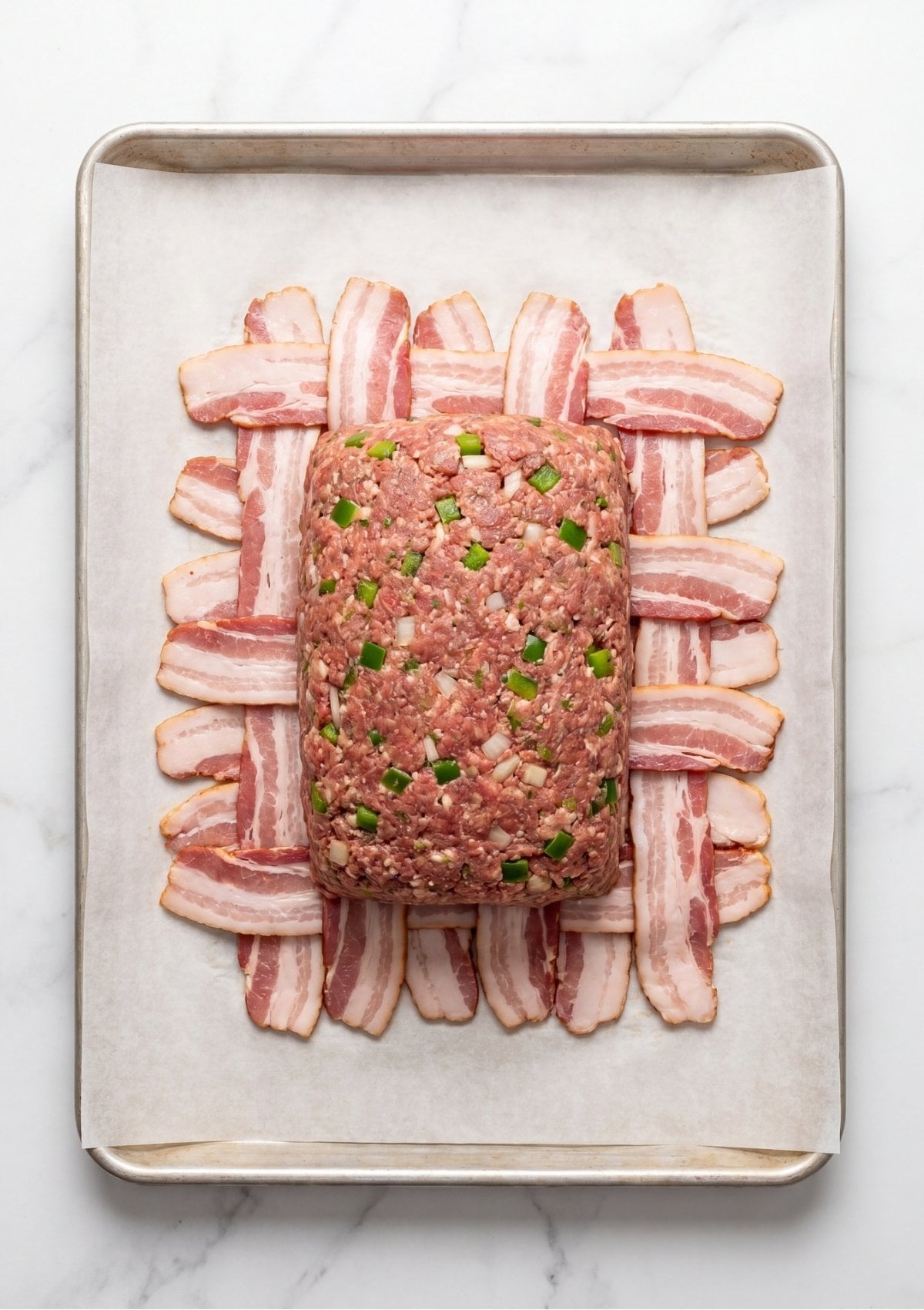 Raw seasoned meatloaf shaped on top of a woven layer of raw bacon strips on a baking sheet