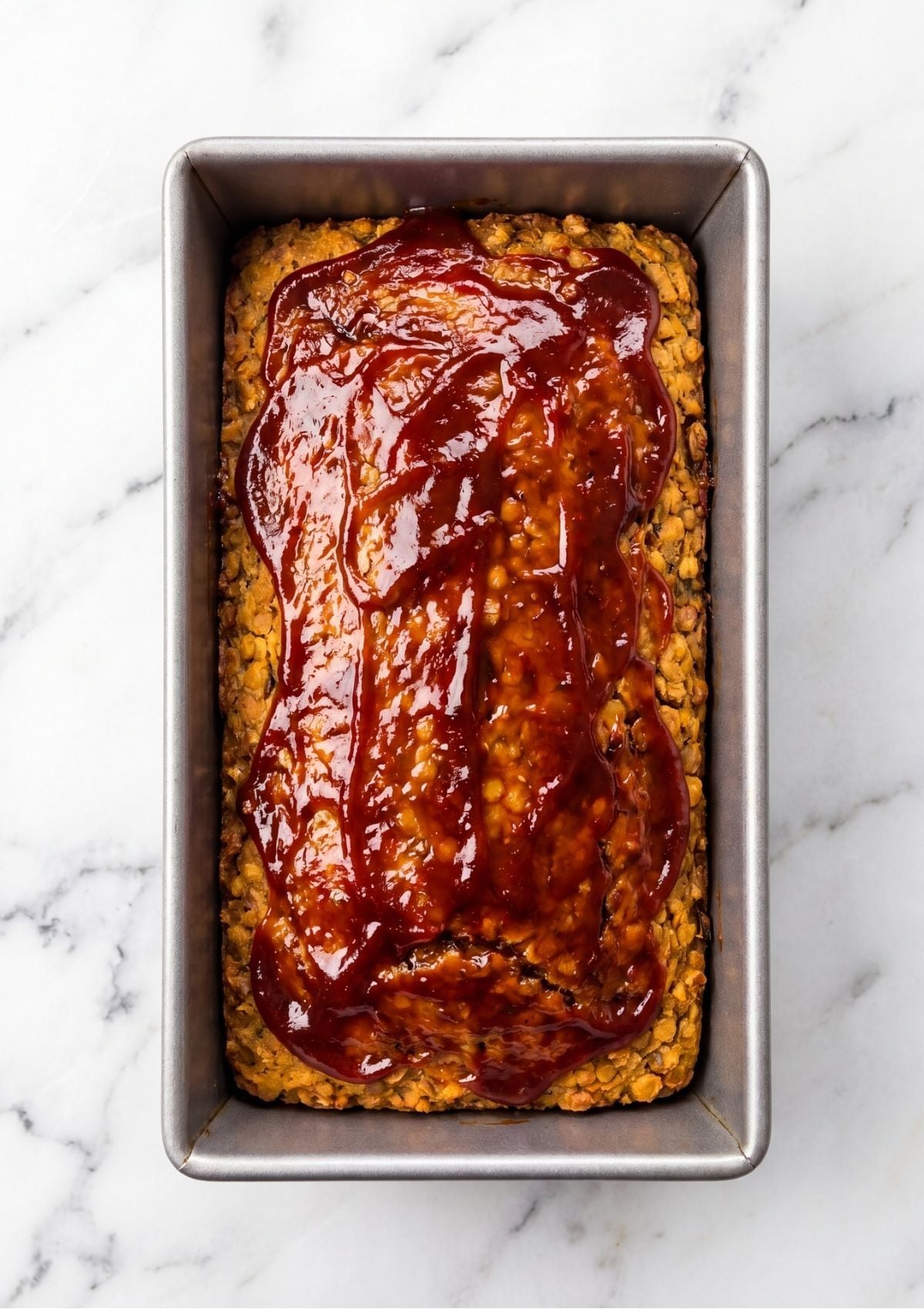 A top down view of a vegan meatloaf in a metal loaf pan topped with a thick red glaze on a marble surface