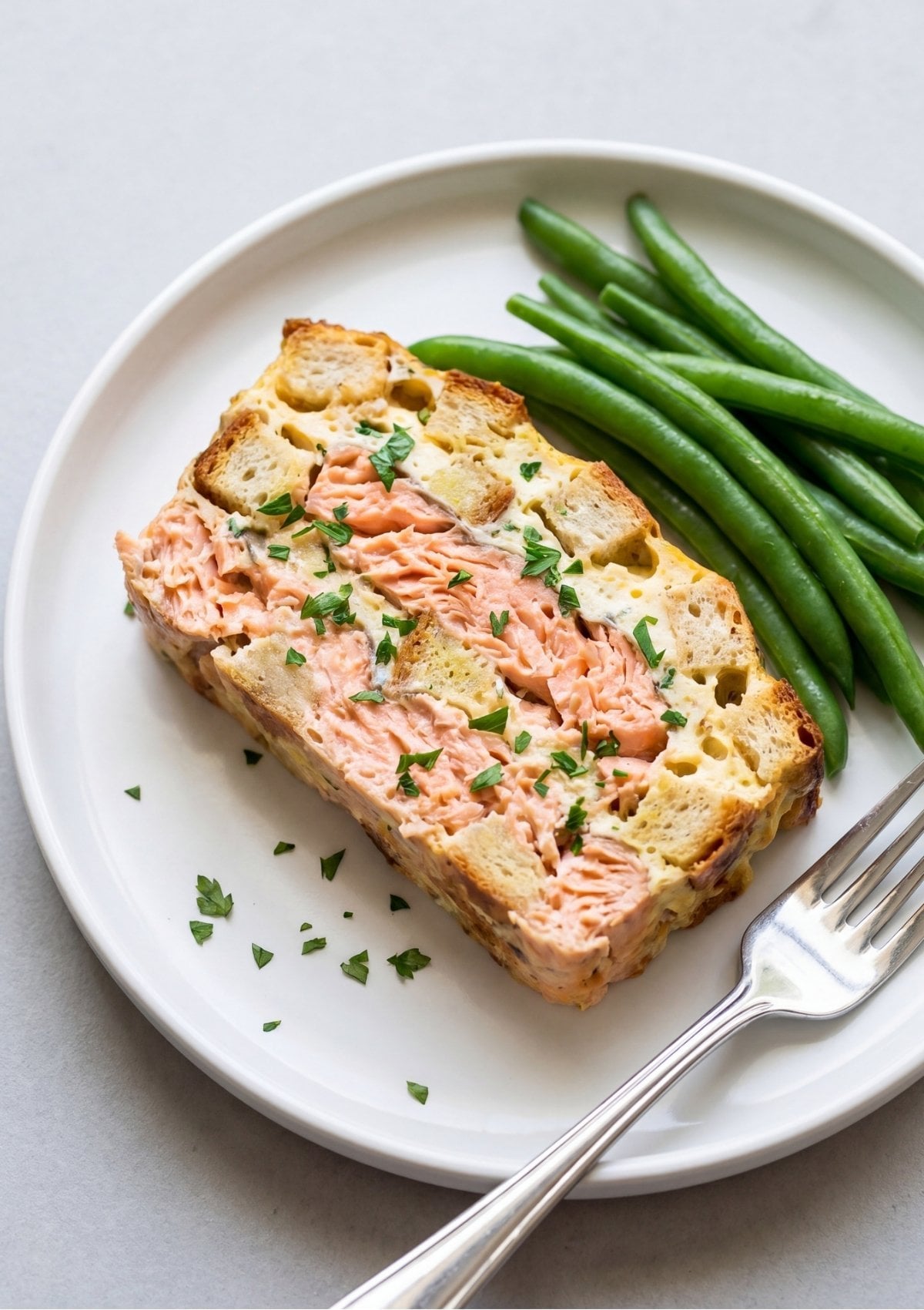 A thick slice of baked salmon meatloaf showing layers of flaked salmon and soft bread served on a white plate with a side of steamed green beans and a fork