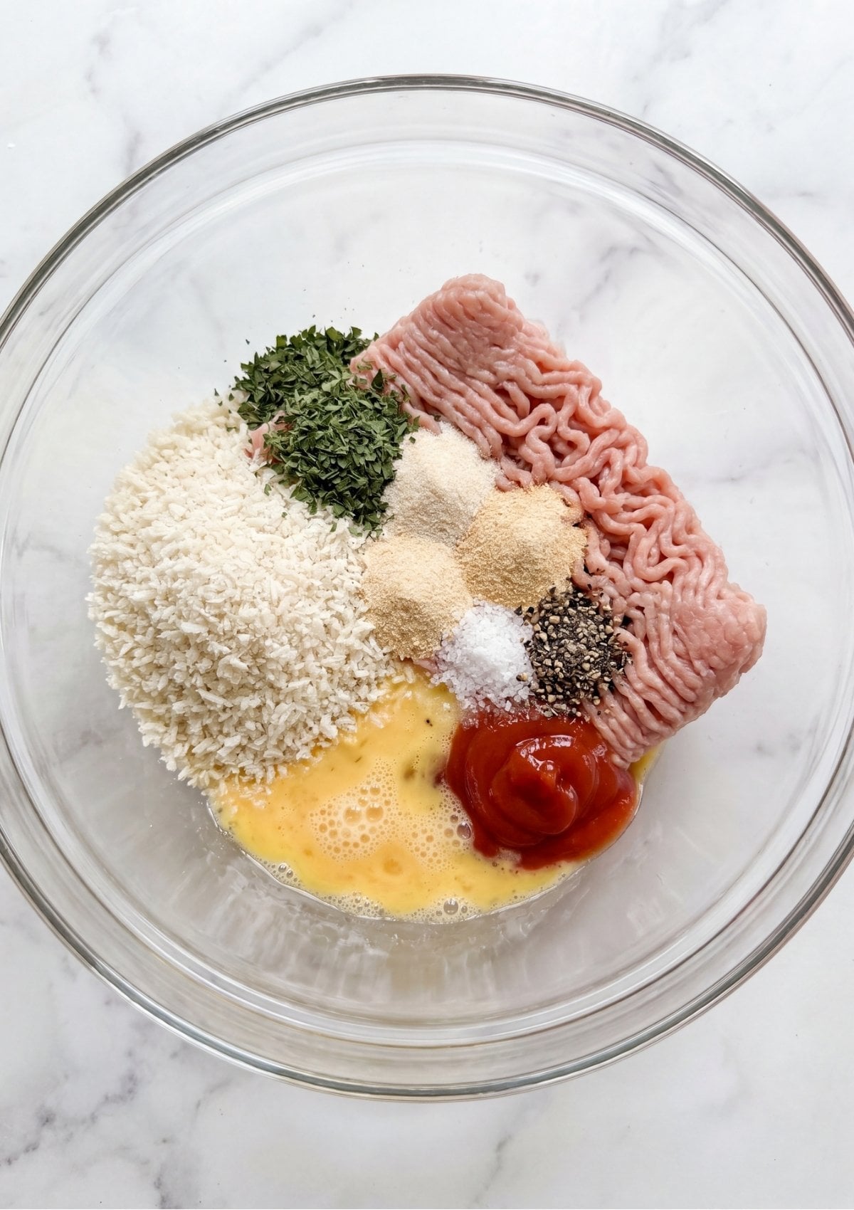 A glass mixing bowl containing ground turkey, breadcrumbs, whisked eggs, ketchup, dried parsley, and various spices.