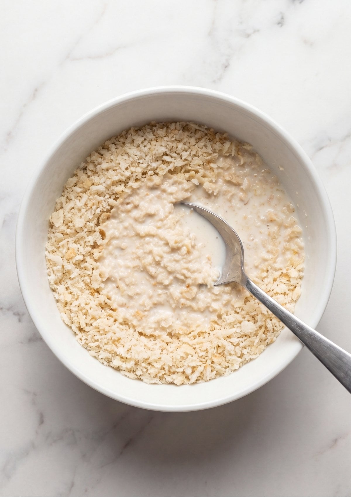 Panko breadcrumbs soaking in a bowl of milk being stirred with a silver spoon on a white marble surface.