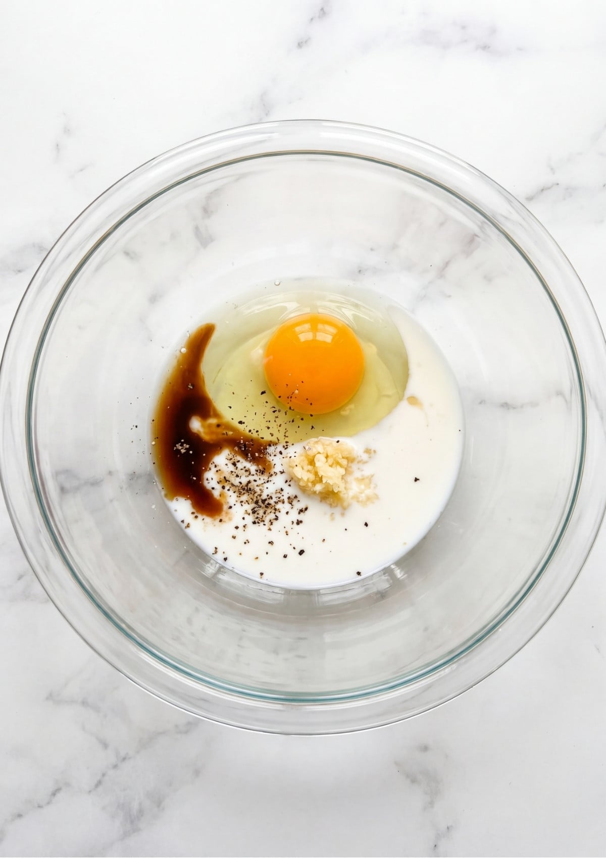 A glass mixing bowl containing a raw egg, milk, minced garlic, Worcestershire sauce, and black pepper on a white marble surface.