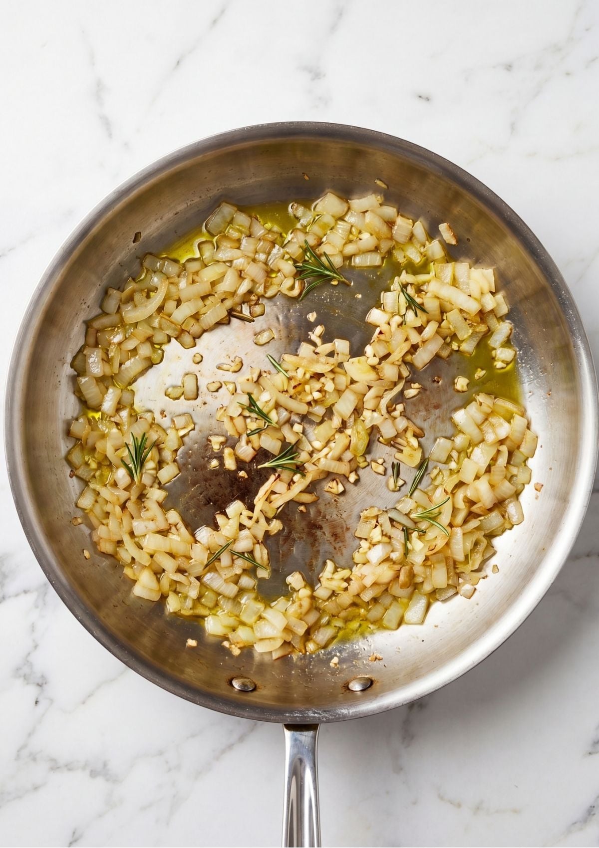 Diced onions, minced garlic, and fresh rosemary sprigs being sautéed in olive oil inside a stainless steel skillet