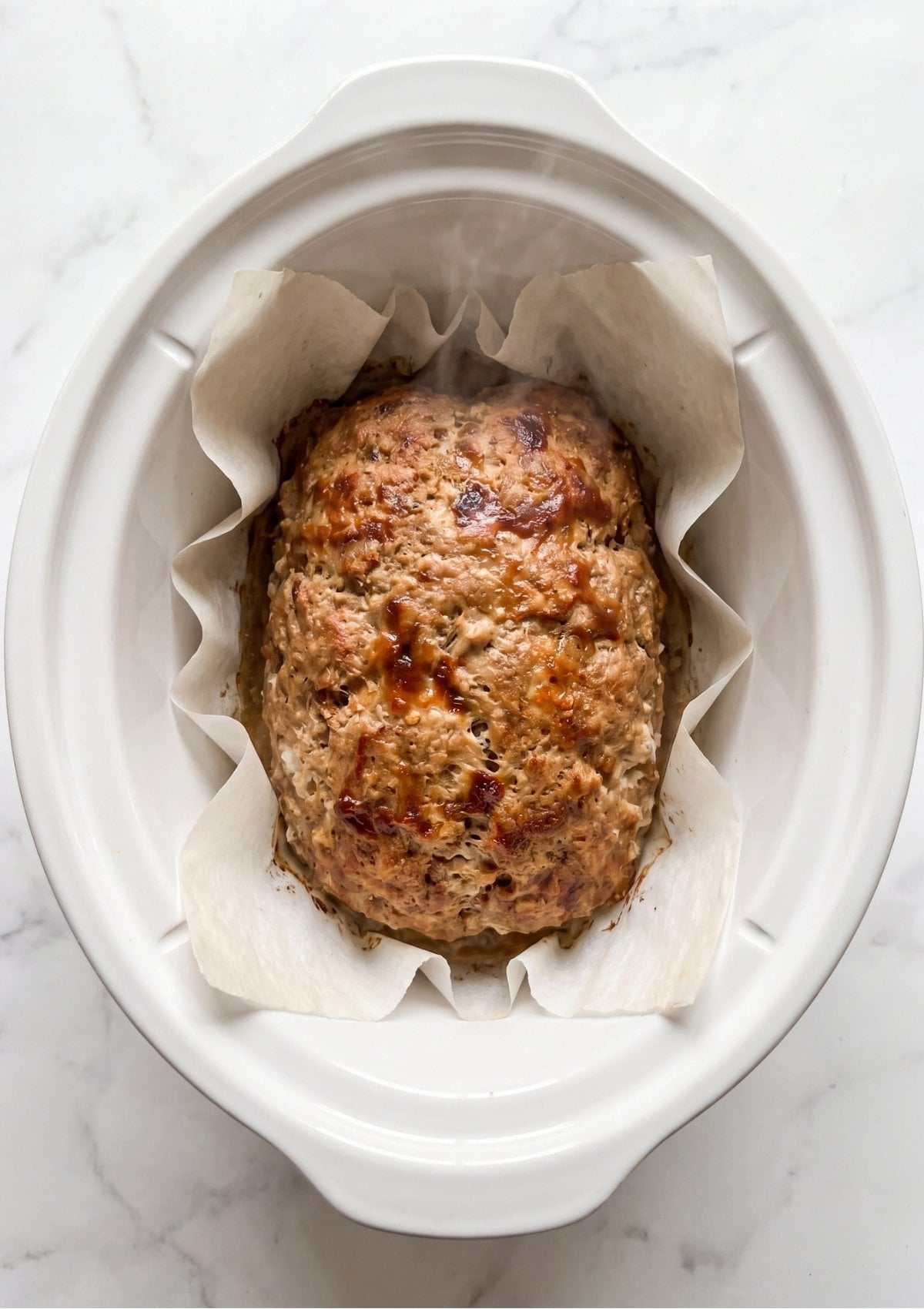 A whole cooked turkey meatloaf steaming inside a white slow cooker lined with parchment paper