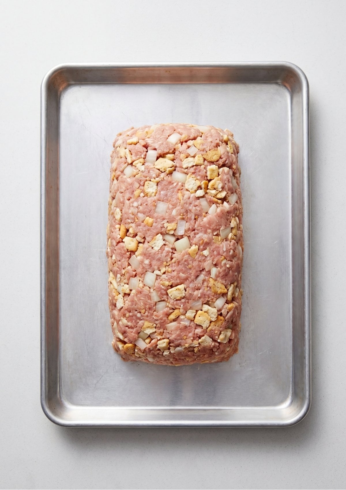 Raw ground turkey meatloaf mixture with chopped onions and cracker pieces shaped into a loaf on a metal baking sheet