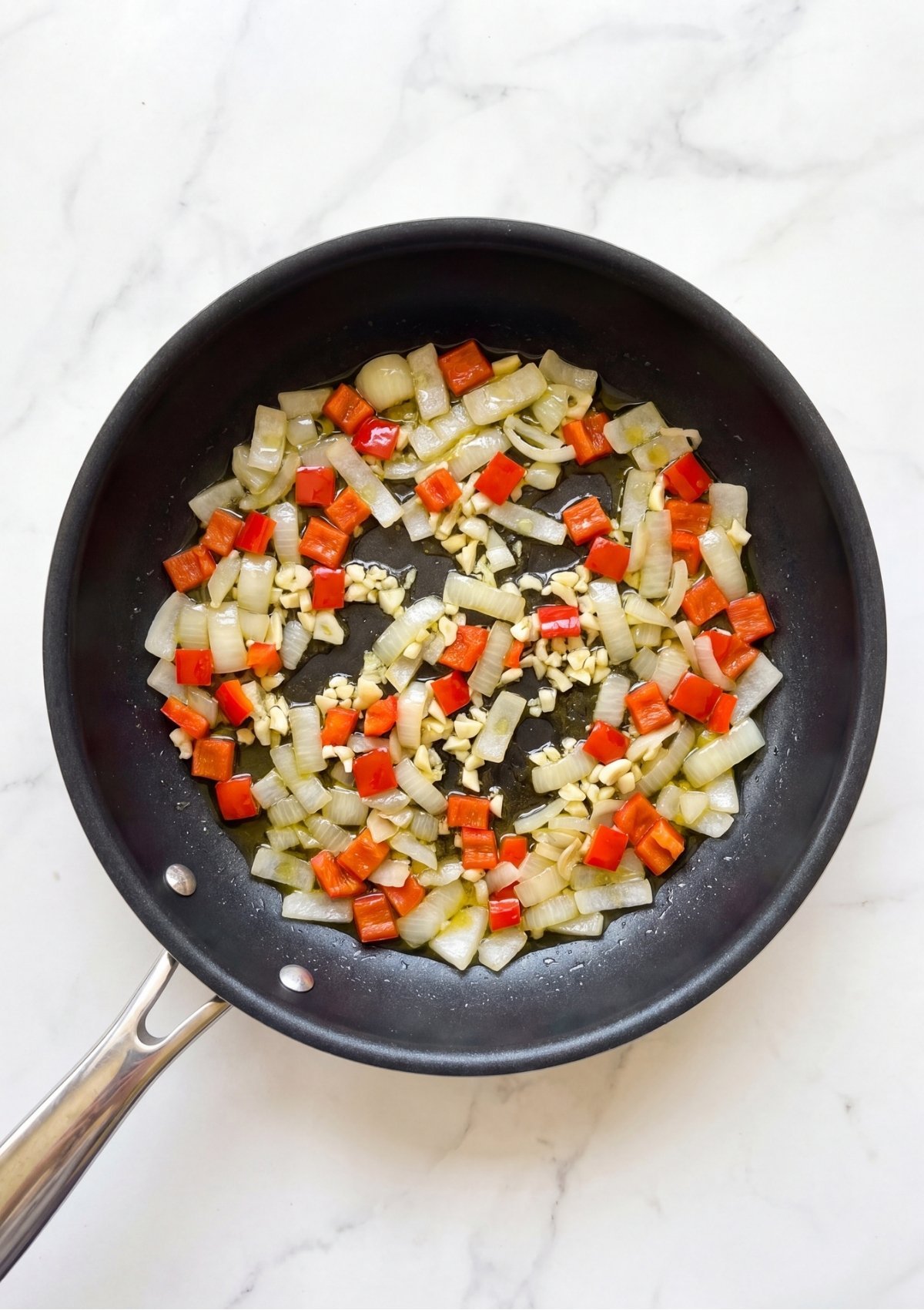 Diced onions, red bell peppers, and minced garlic cooking with olive oil in a black skillet placed on a white marble countertop