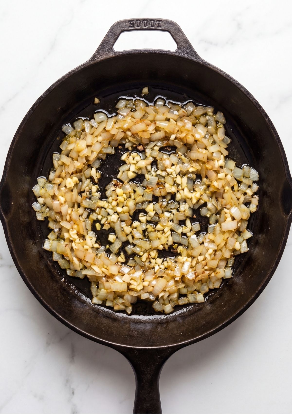 Finely diced onions and minced garlic cooking in olive oil inside a black cast iron skillet.
