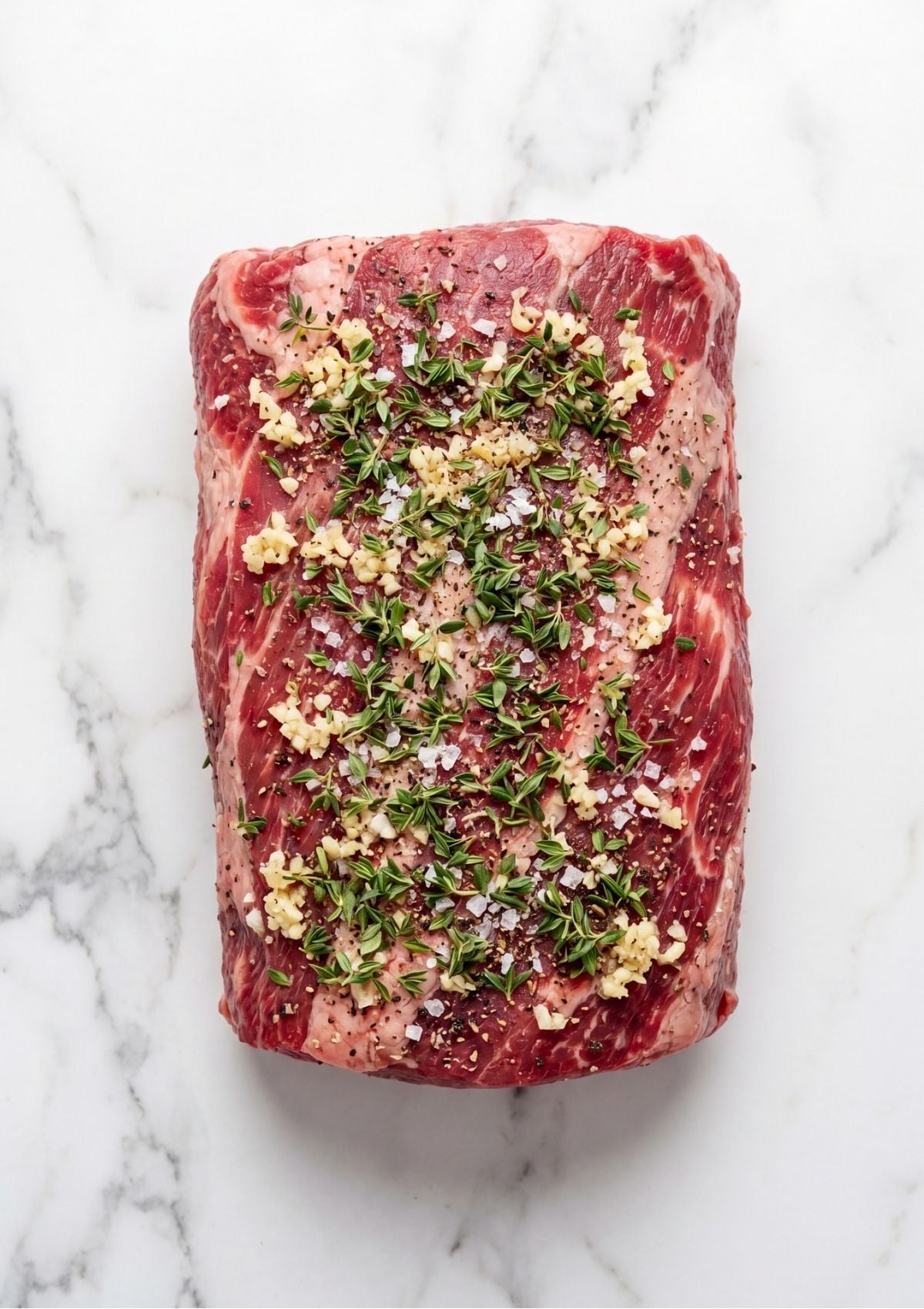 Overhead view of a raw boneless beef top loin roast seasoned evenly with minced garlic, fresh thyme leaves, coarse salt, and black pepper