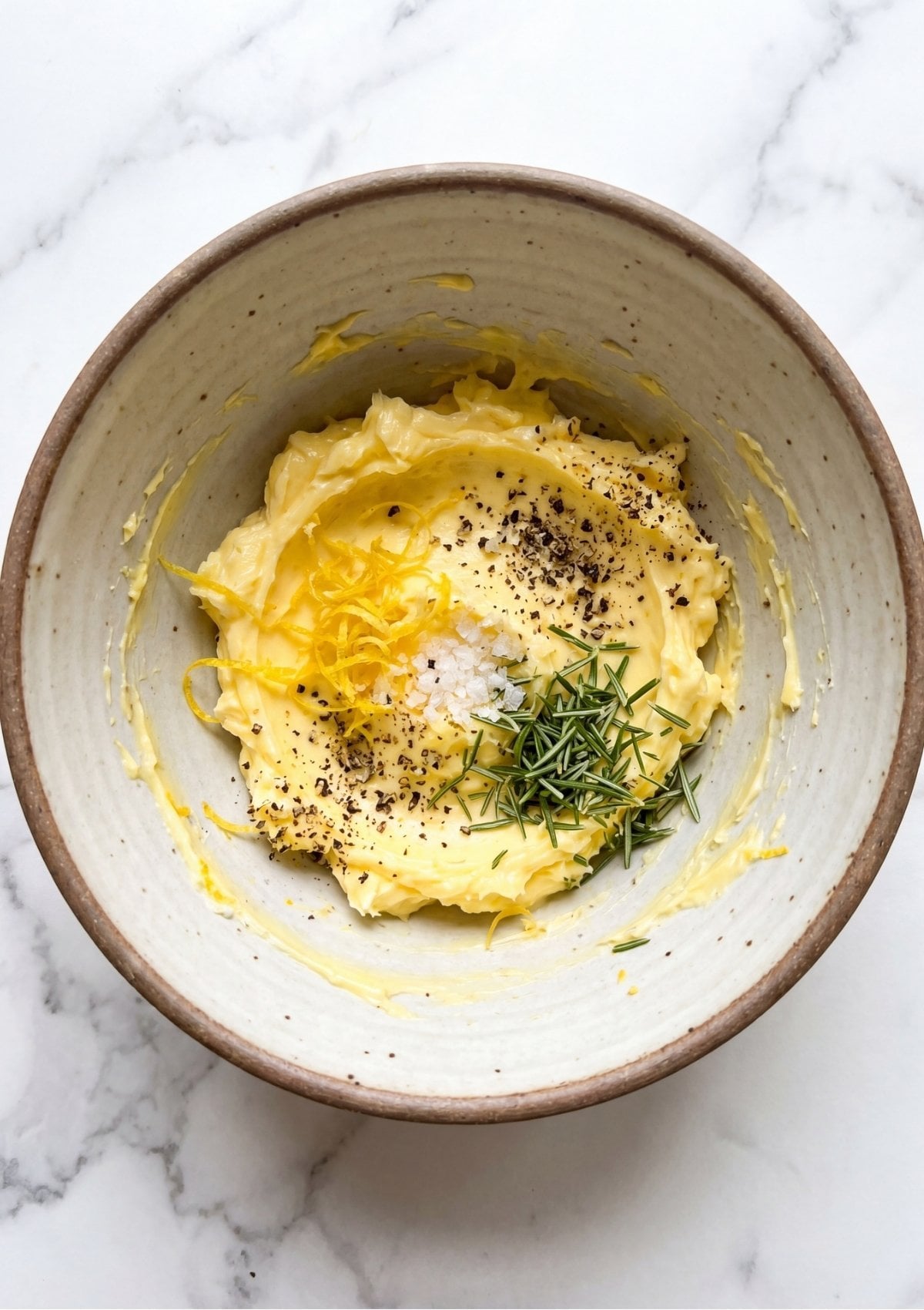 Overhead view of a ceramic bowl containing softened yellow butter topped with fresh lemon zest, cracked black pepper, sea salt, and chopped rosemary