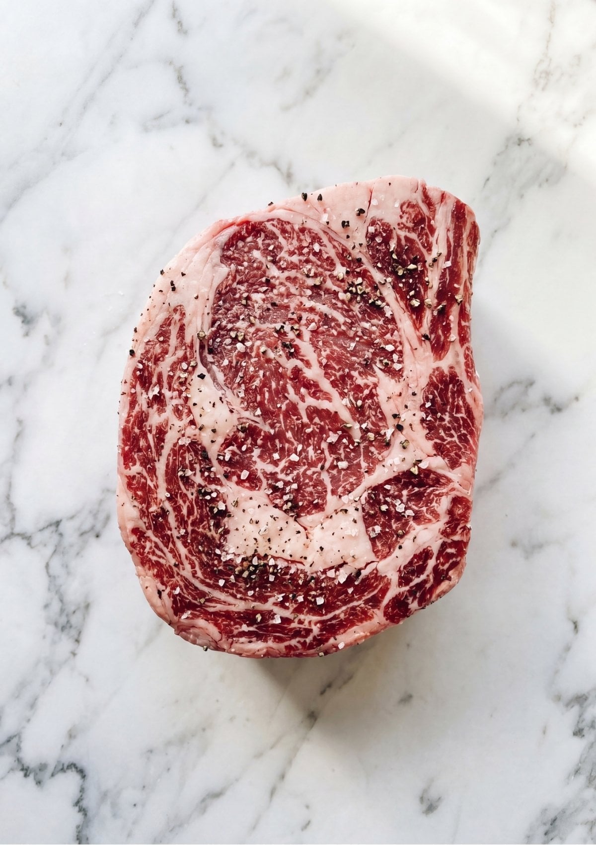 A raw marbled beef arm roast seasoned generously with coarse salt and cracked black pepper on a white marble background.