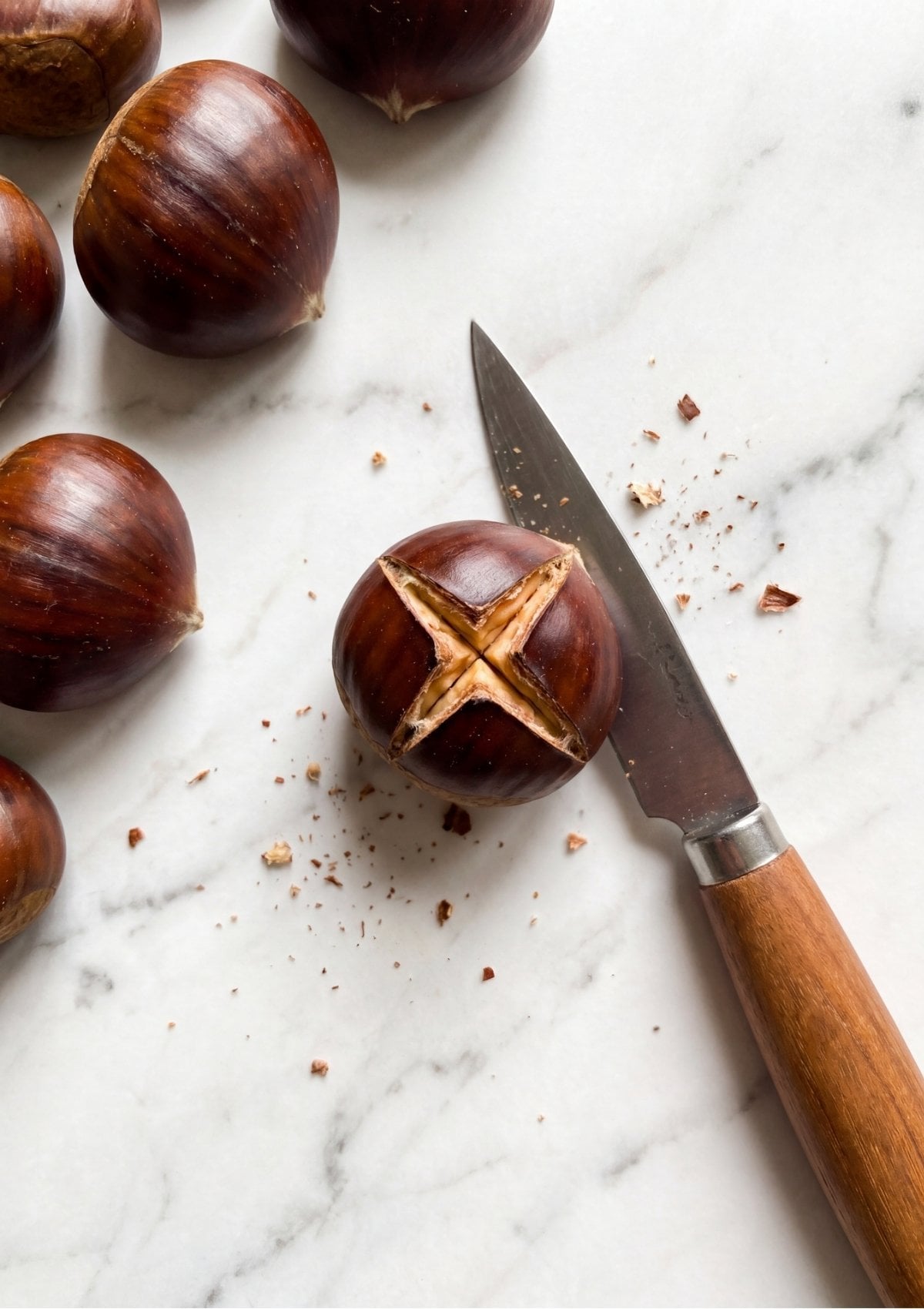A sharp paring knife with a wooden handle resting next to a whole chestnut that has a deep X scored into its brown shell.