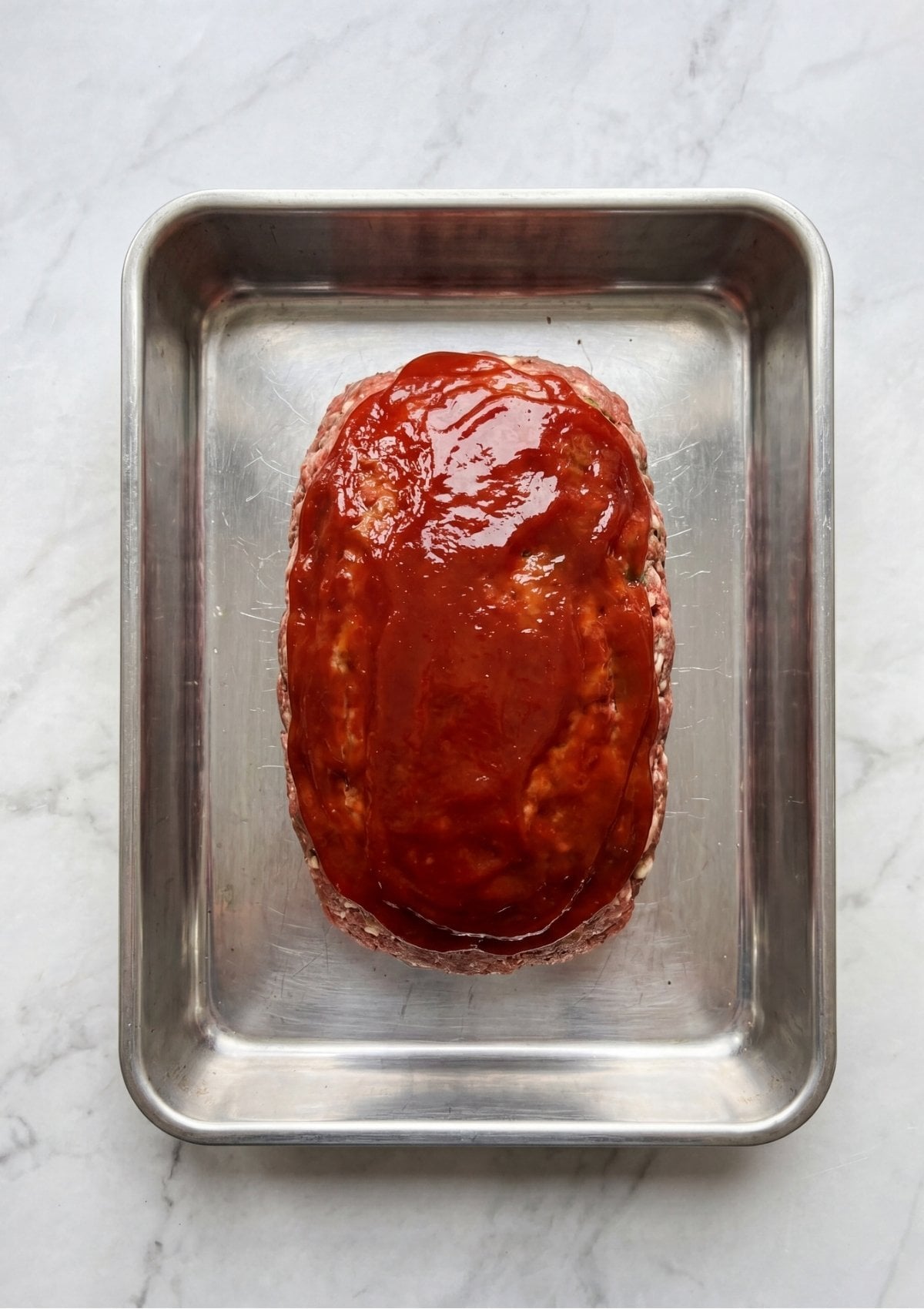Raw ground beef shaped into a loaf and topped with a smooth layer of red glaze in a metal baking dish.