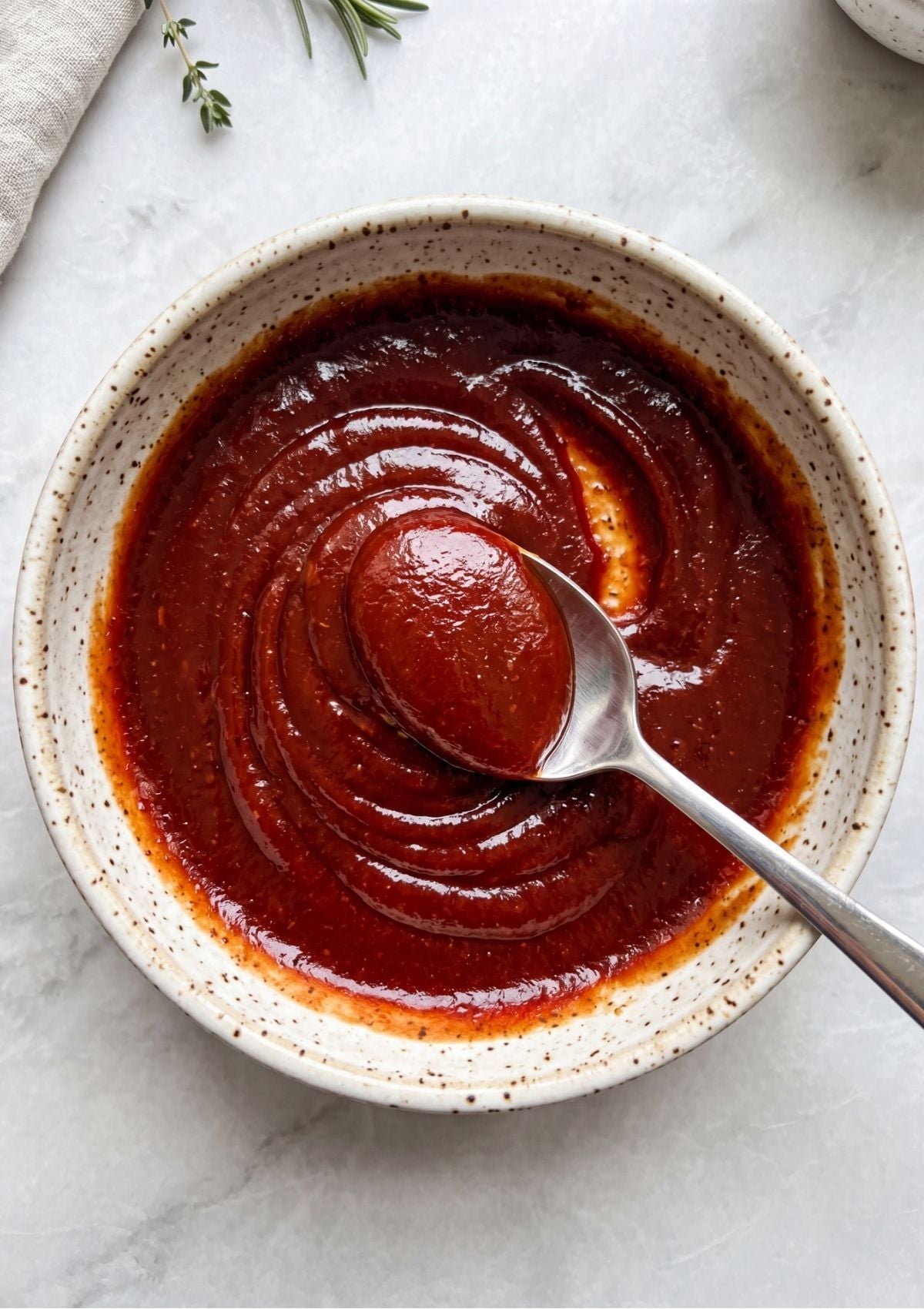 A silver spoon lifting a dollop of smooth, glossy Meatloaf Glaze Recipe from a speckled bowl.