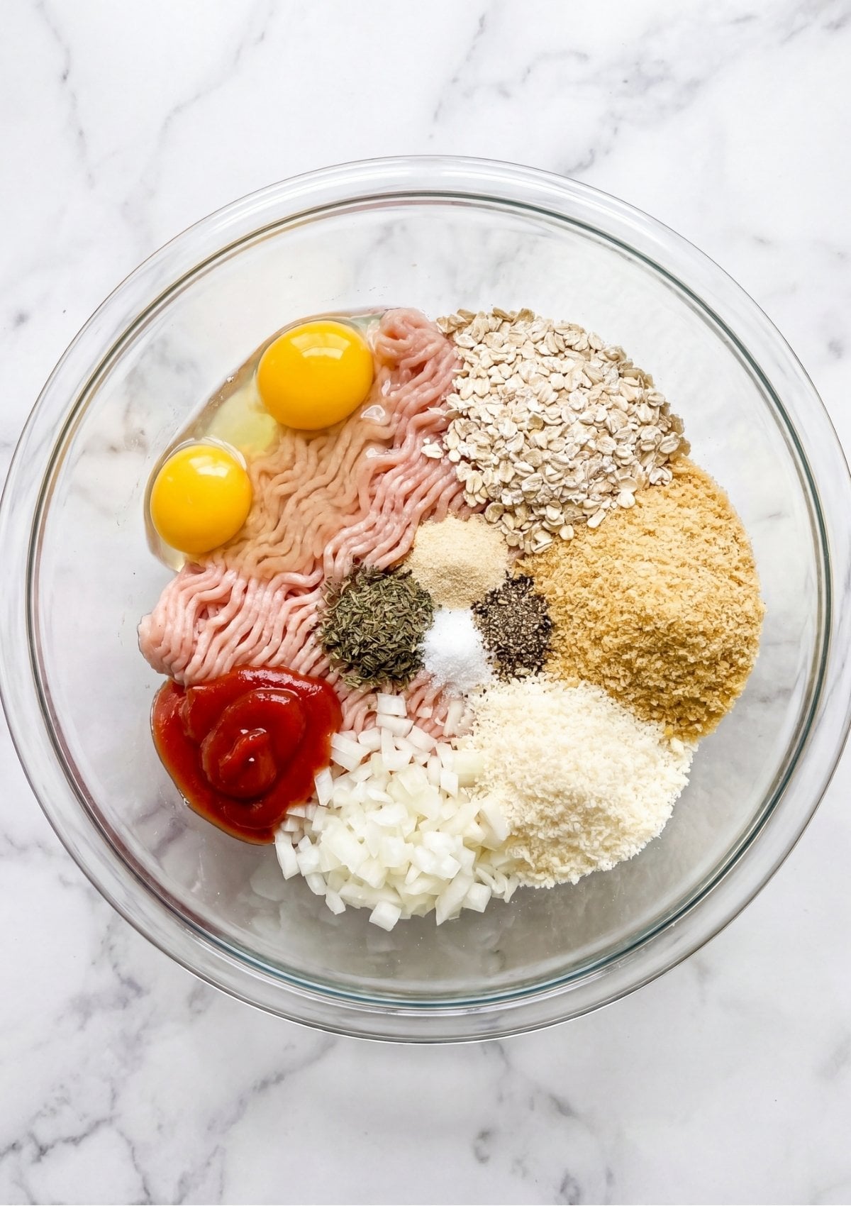 A glass mixing bowl filled with raw ground chicken, two eggs, oats, breadcrumbs, diced onions, ketchup, and various dried herbs and spices.
