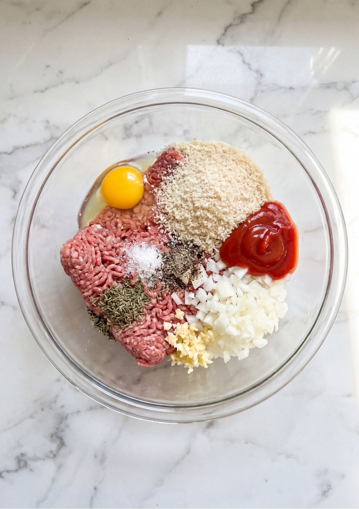 A clear glass bowl showing separated ingredients before mixing: ground beef, a whole egg, breadcrumbs, ketchup, diced onions, garlic, and spices.